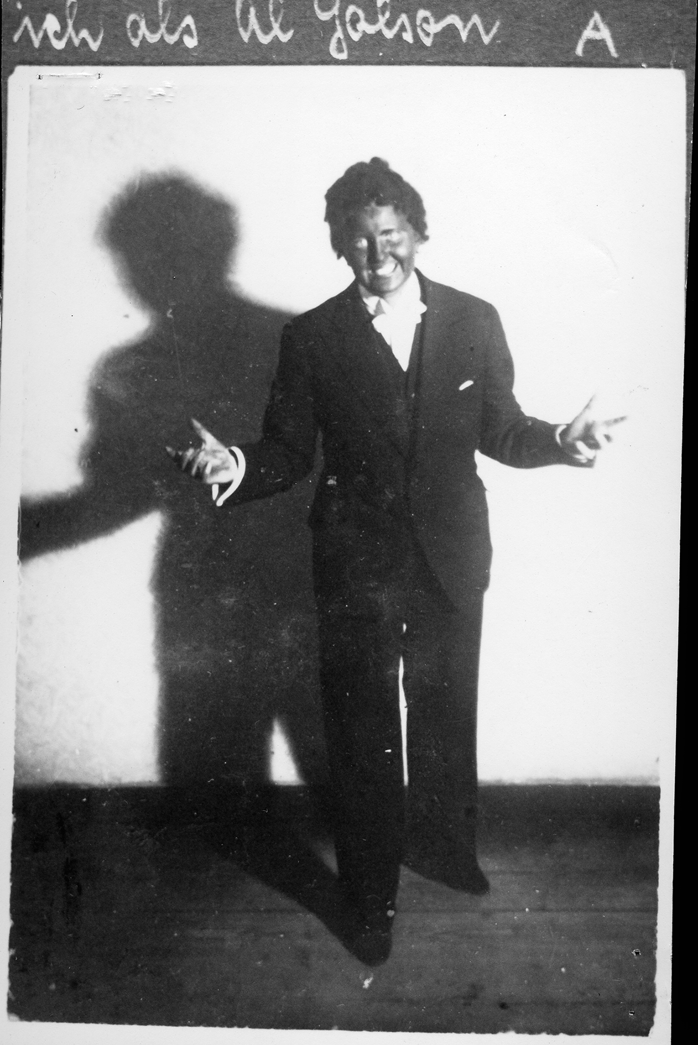 Ein Schwarzweißfoto zeigt eine lachende Person in Blackface, die in einem dunklen Anzug mit ausgebreiteten Armen vor einer weißen Wand posiert. Auf der Wand ist ihr Schatten zu sehen.