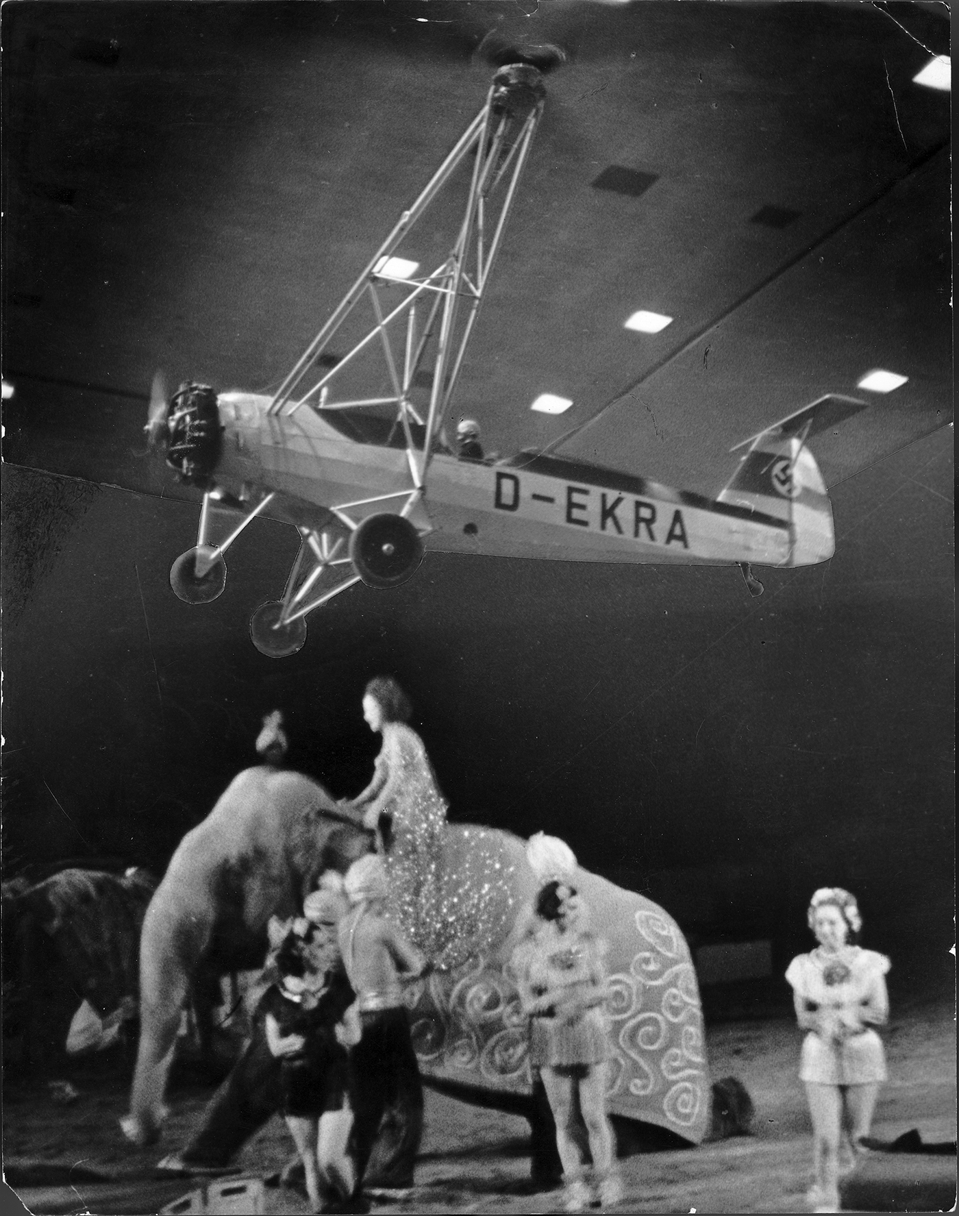 Ein Schwarzweißfoto zeigt einen Focke-Hubschrauber, der durch eine schwarze, beleuchtete Halle fliegt. Darunter sind ein Elefant und mehrere kostümierte Personen zu sehen.