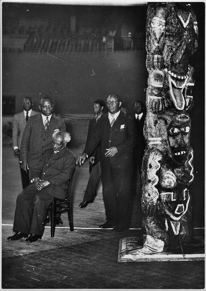 Ein schwarzweißfoto zeigt sechs Personen in Männeranzügen, die neben einem Totempfahl in einer Halle stehen.