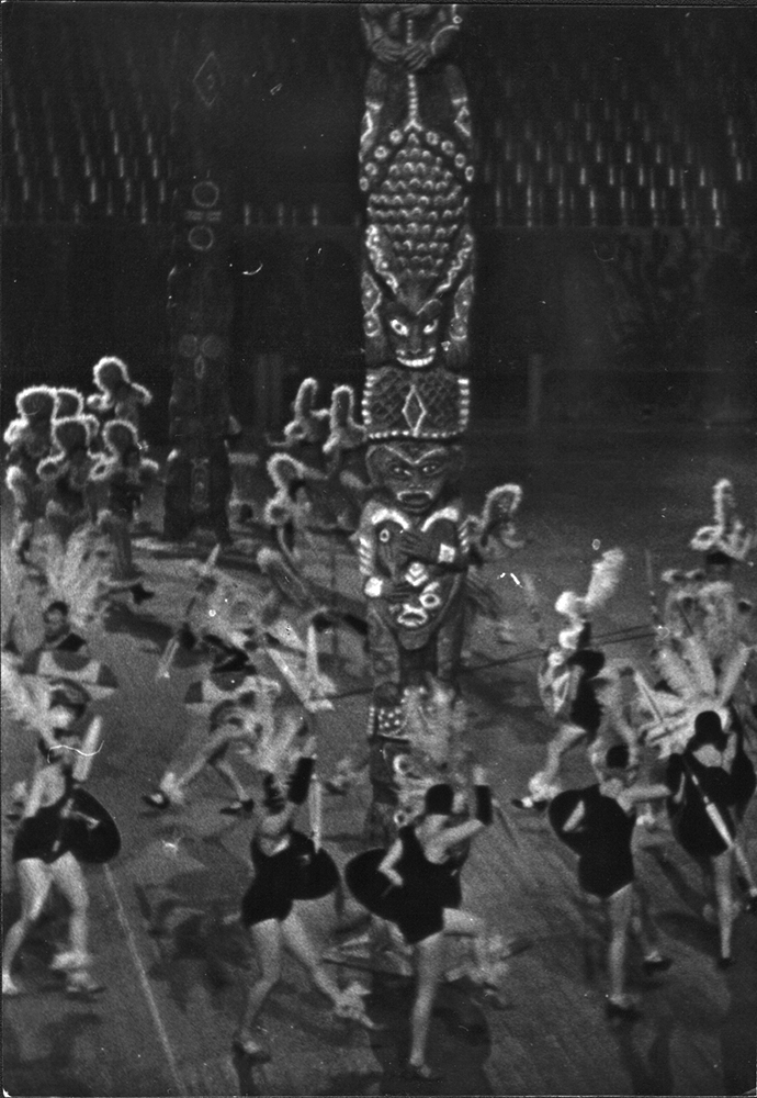 Ein Schwarzweißfoto zeigt Revue Girls in Federkostümen, die in einer Halle um zwei Totempfähle tanzen.