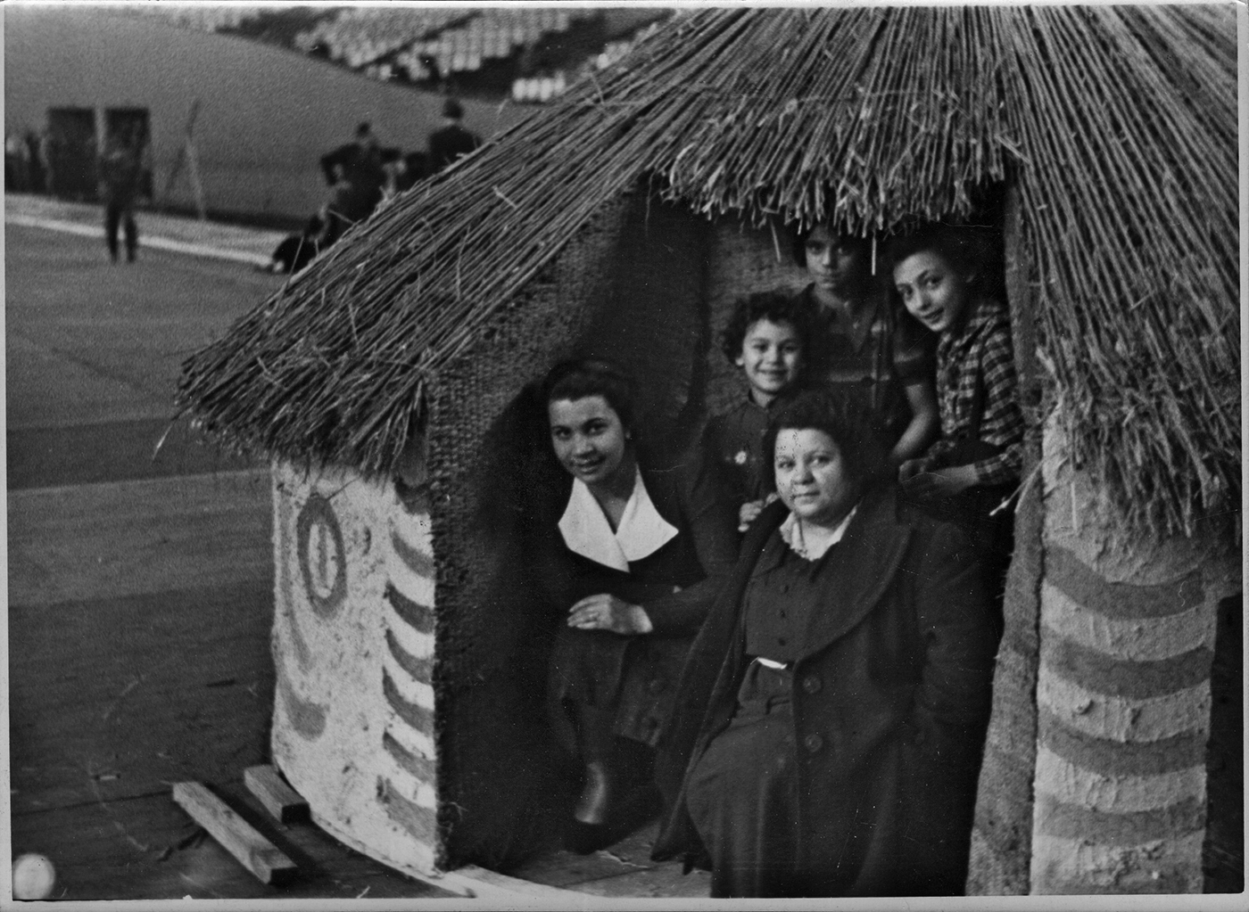 Ein Schwarzweißfoto zeigt eine Gruppe von fünf Personen, die in einer kleinen, runden Hütte mit Bastdach sitzen. Im Hintergrund ist eine Halle zu erkennen.