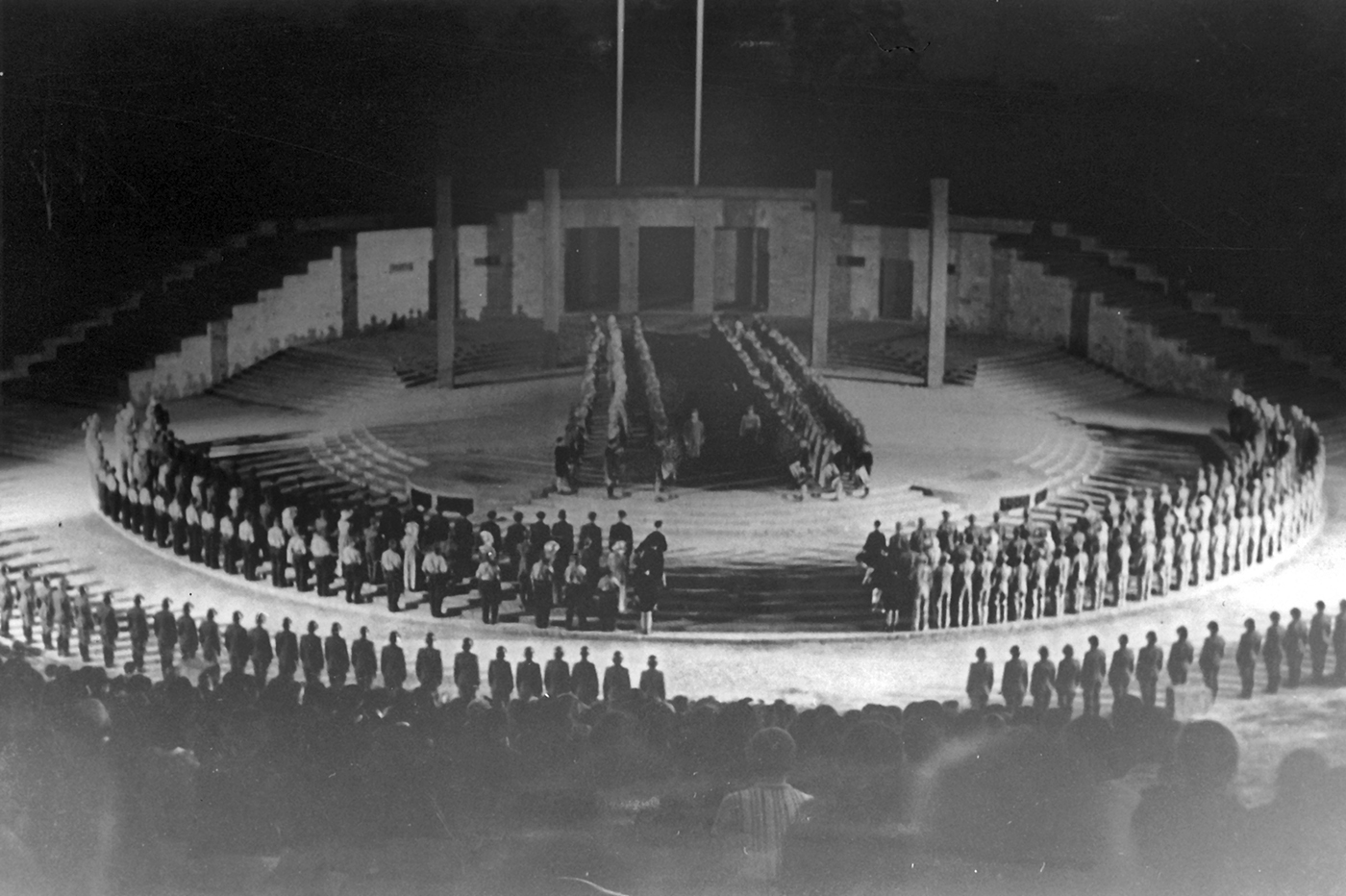 Ein Schwarzweißfoto zeigt eine runde Freilichtbühne, auf der, angeordnet in zwei äußeren Kreisen und zwei zentrierten Reihen, Gruppen von Darstellenden in Uniform zu sehen sind.