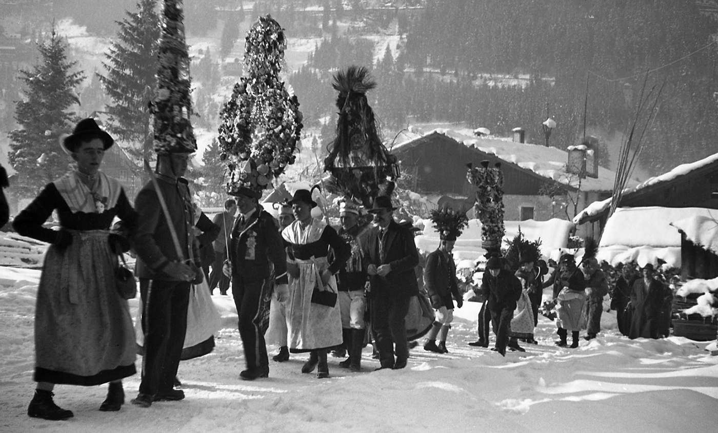 Ein Schwarzweißfoto zeigt einen Perchtenzug auf einem schneebedeckten Hang.