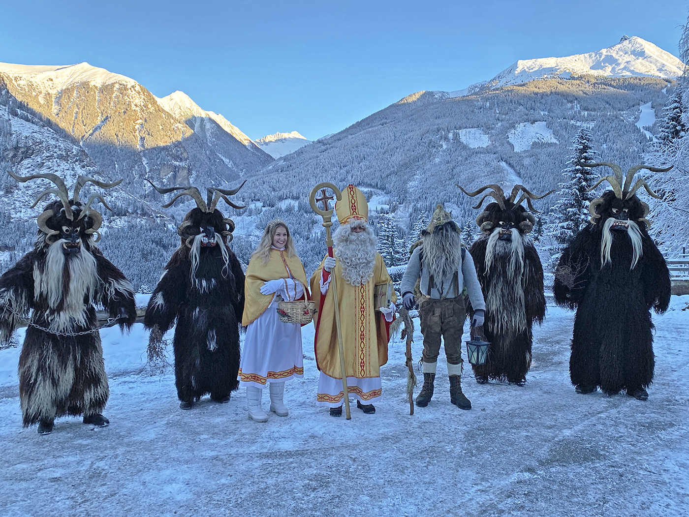 Ein Farbfoto zeigt sieben Personen in traditionellen Krampus- und Nikolauskostümen, die vor schneebedeckten Bergen stehen.