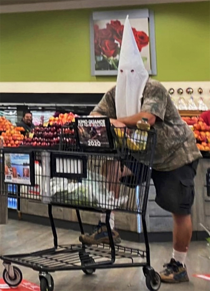 A color photo shows a person wearing a white Ku Klux Klan mask, leaning on a shopping cart in a supermarket.