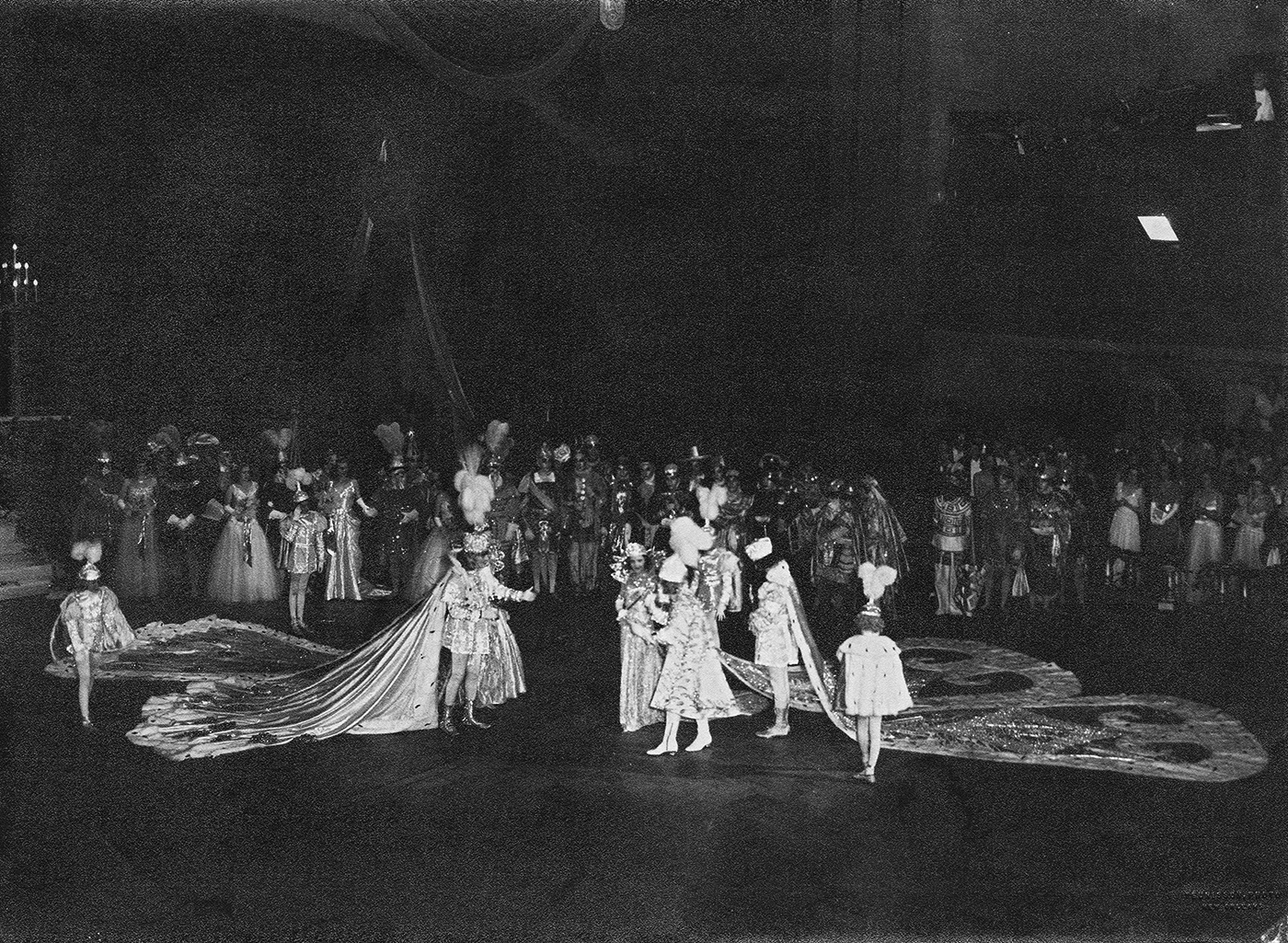 A black-and-white photo shows many people in a large hall. In the center, two figures in opulent royal costumes with meter-long trains meet; the figure on the right is accompanied by four others.