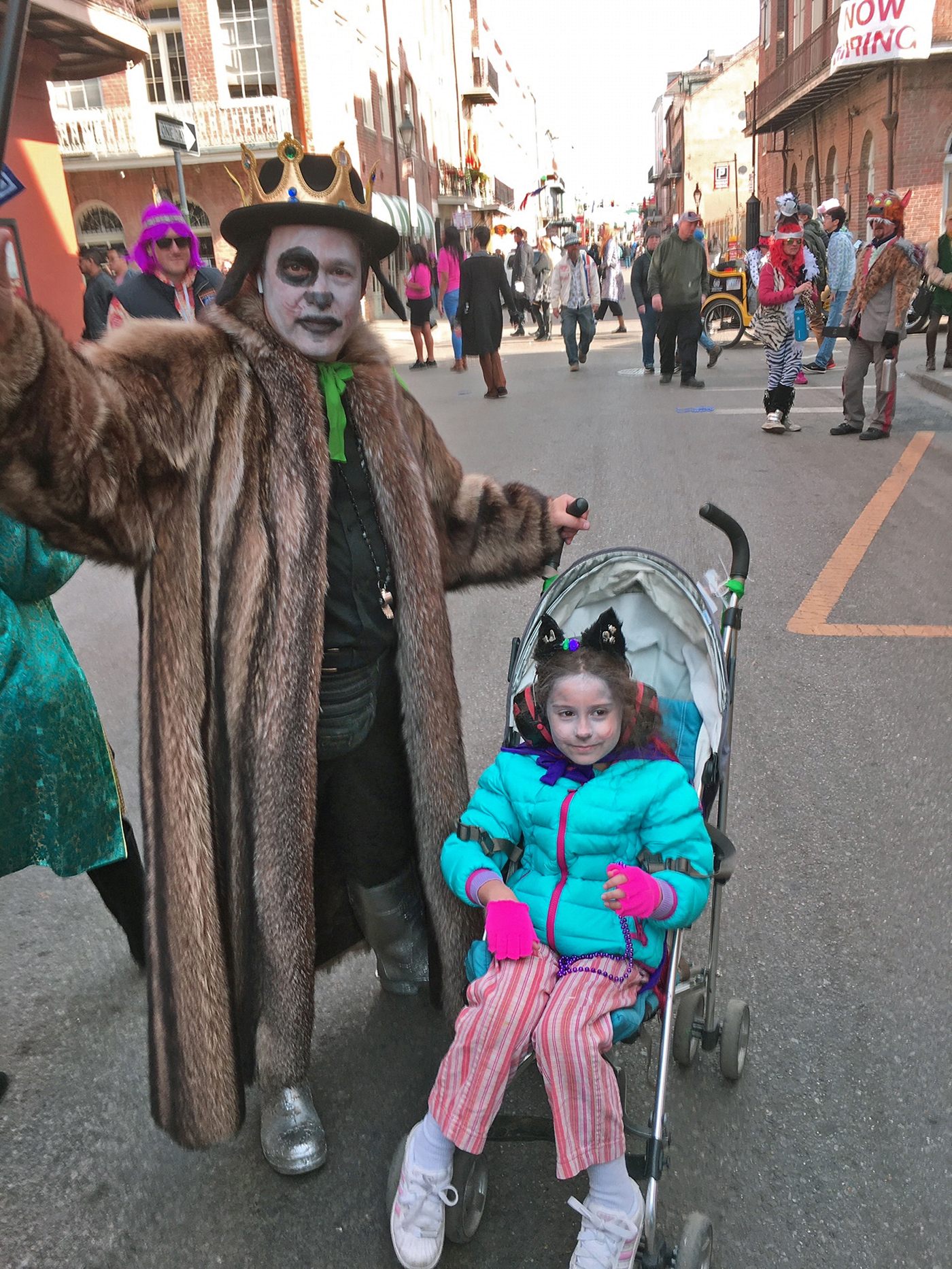 Ein Farbfoto zeigt eine Person in Pelzmantel und mit Krone, das Gesicht weiß, ein Auge schwarz geschminkt, die mit einem ebenfalls verkleideten Kind, das in einem Kinderwagen sitzt, lachend vor der Kamera posiert.