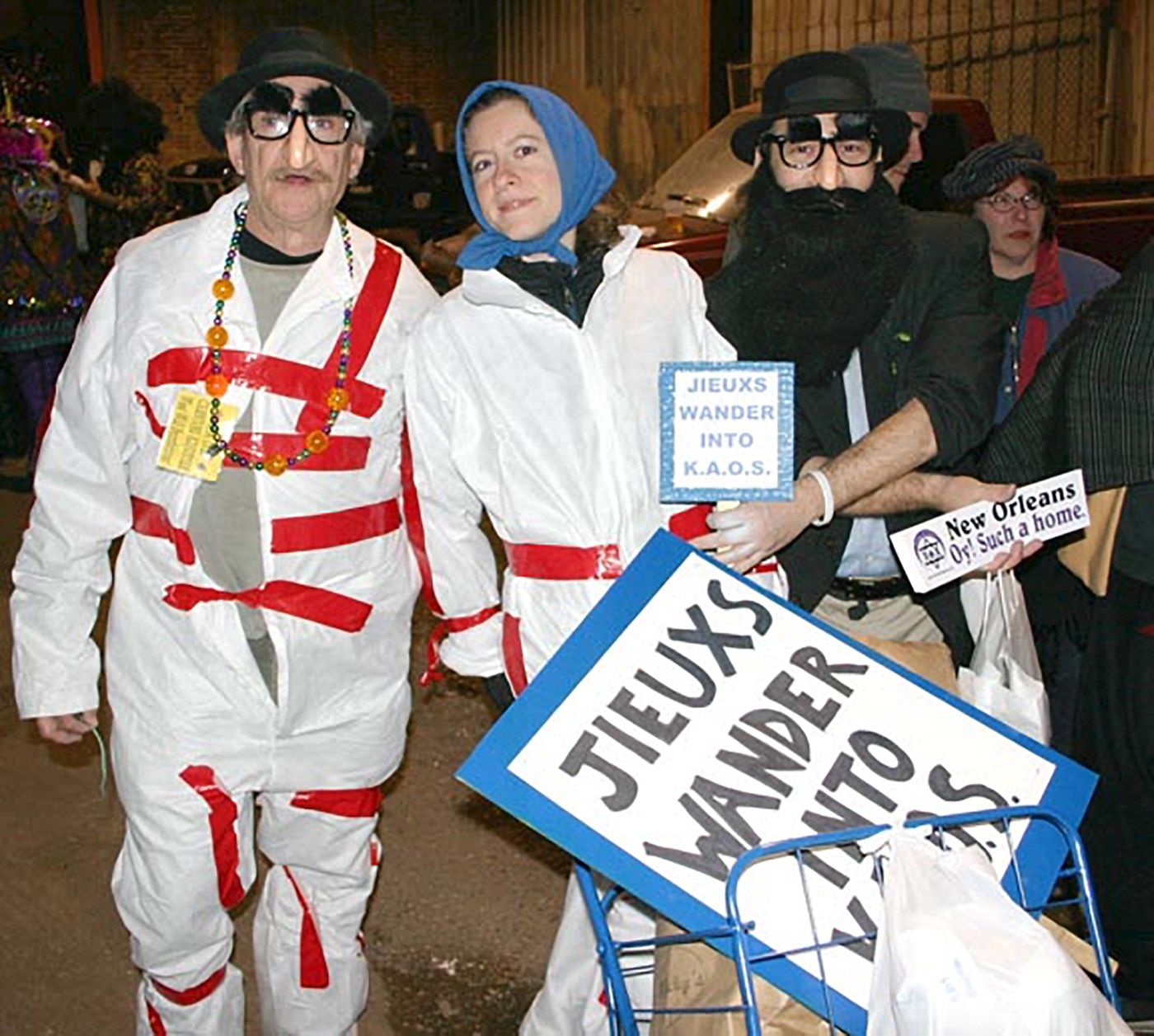 Ein Farbfoto zeigt drei kostümierte Personen in weißen Schutzanzügen, auf denen rote Klebebänder angebracht sind. Zwei tragen Scherzbrillen mit künstlichen Augenbrauen und Nasen und schwarze Hüte, eine weitere Person ein blaues Kopftuch. Sie halten Schilder mit der Aufschrift „Jieux wander into K.A.O.S.“