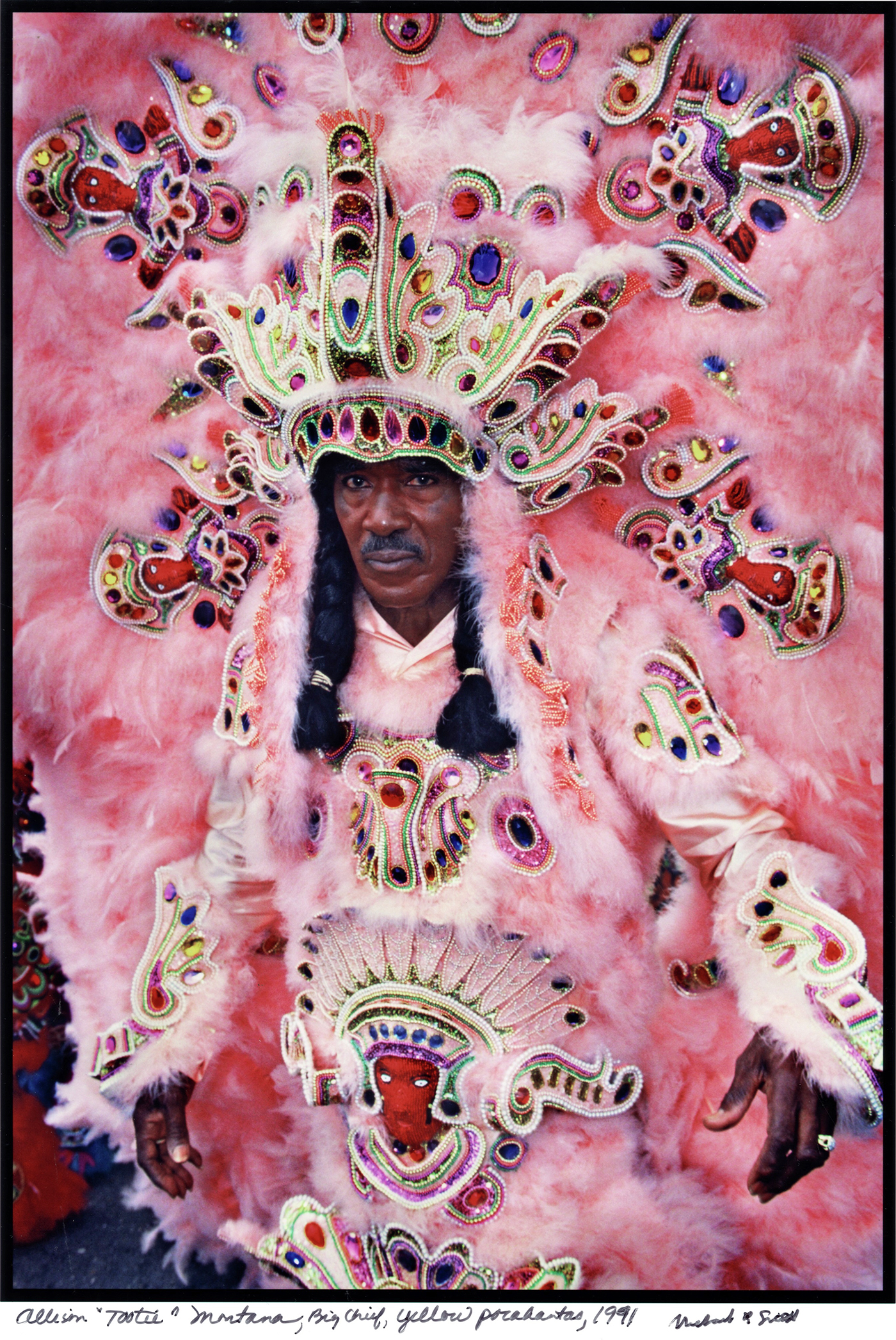 Ein Farbfoto zeigt einen Mann mit zwei schwarzen Zöpfen in einem überkörpergroßen, rosafarbenen Federkleid mit bunten Perlenstickereien.