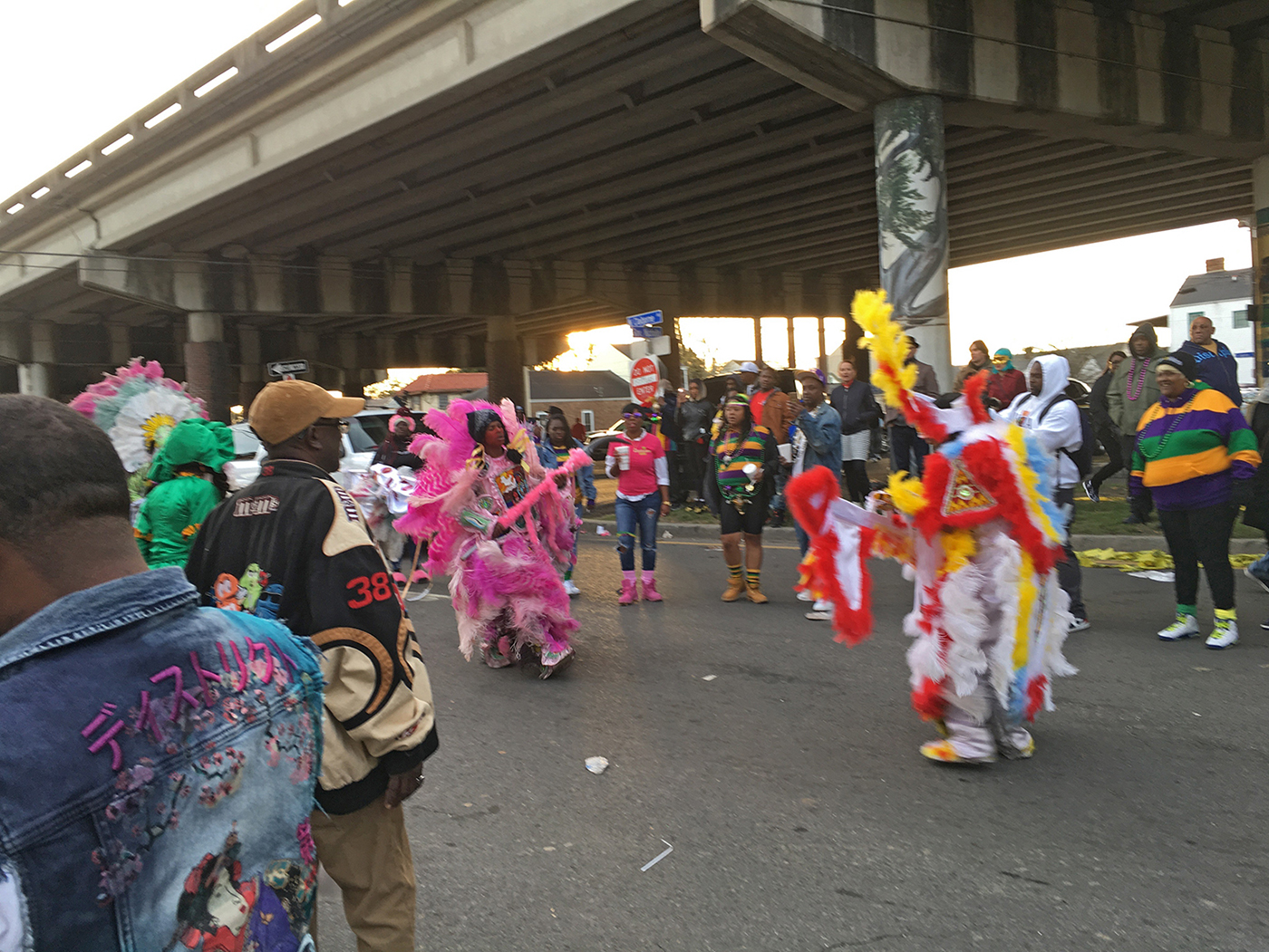 Ein Farbfoto zeigt zwei Personen in bunten Federkleidern, die sich auf einer Straße unter einer Überführung konfrontieren und von den Umstehenden beobachtet werden.