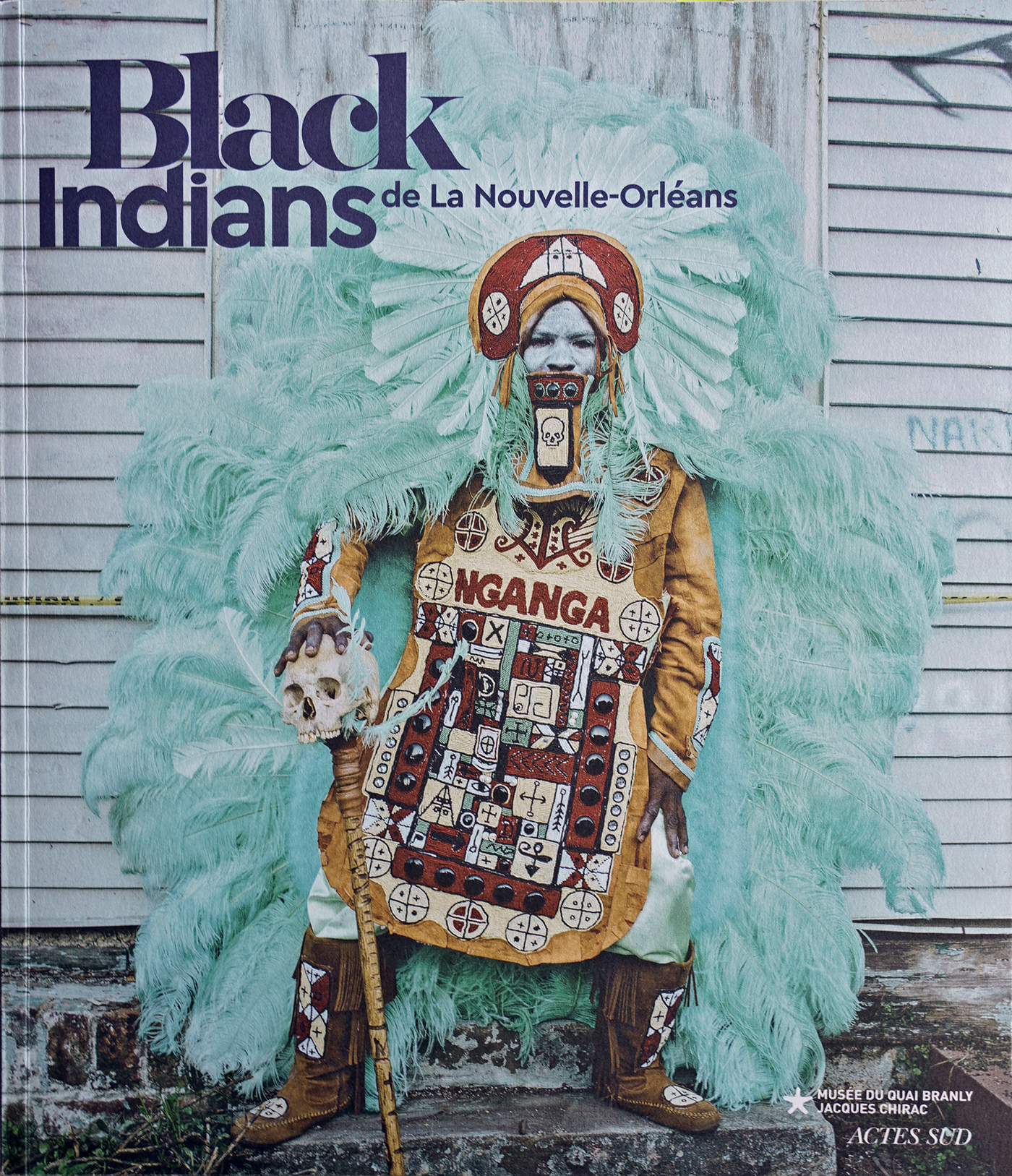 Ein farbiges Katalogcover von Black Indians de la Nouvelle-Orléans zeigt eine weiß geschminkte Person in einem türkisfarbenen, überkörpergroßen Federkleid breitbeinig posierend. Sie hält einen Stab mit Schädelspitze.