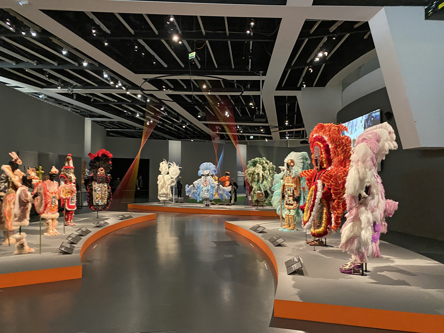 Ein Farbfoto zeigt einen Ausstellungsraum, in dem kunstvolle, farbenfrohe Federkleidskulpturen in Rot-, Rosa-, Blau- und Orangetönen ausgestellt sind.