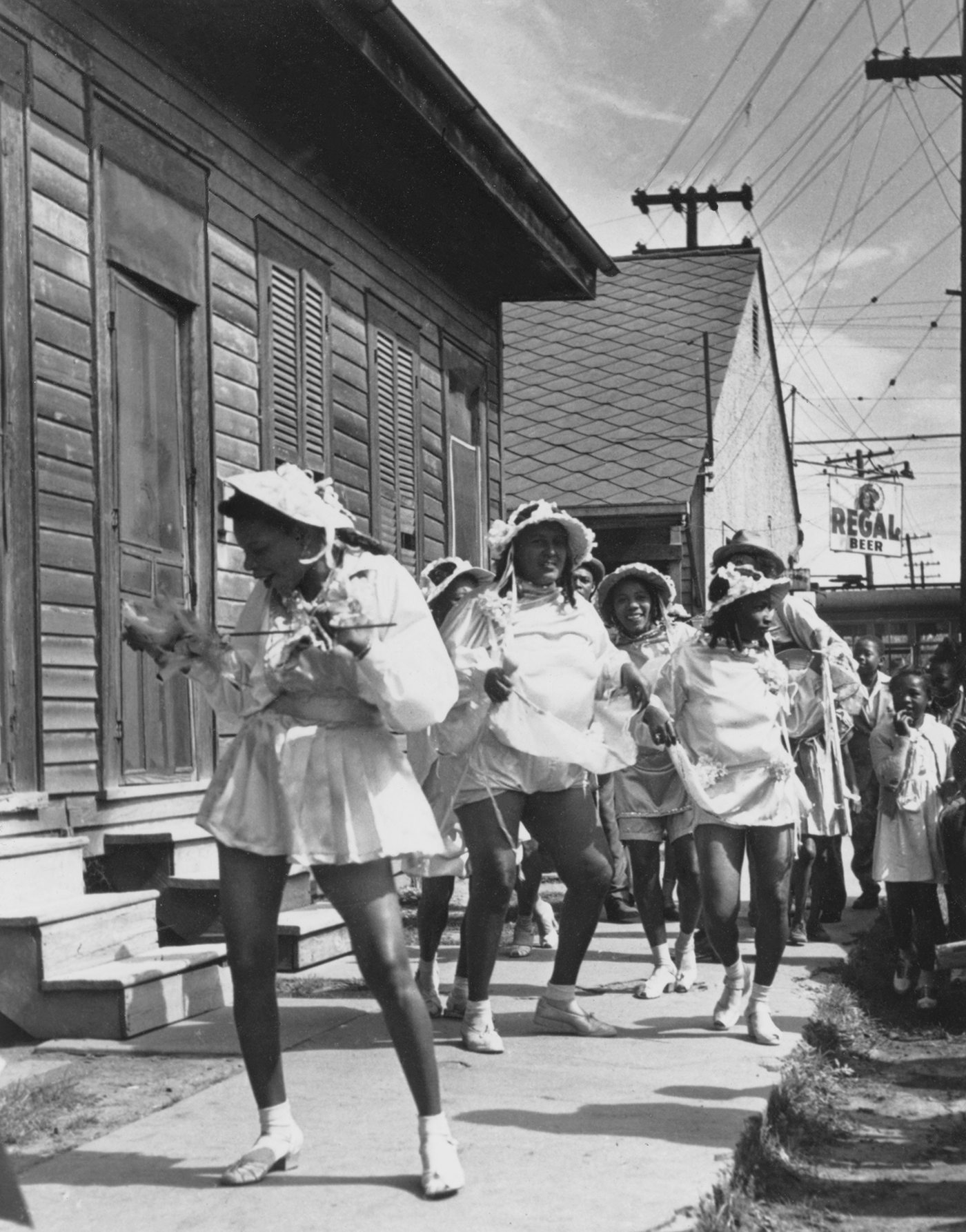 Ein Schwarzweißfoto zeigt eine tanzende Gruppe in kurzen weißen Kleidern und mit berüschten Hüten neben einem Holzgebäude. Stromleitungen und ein 