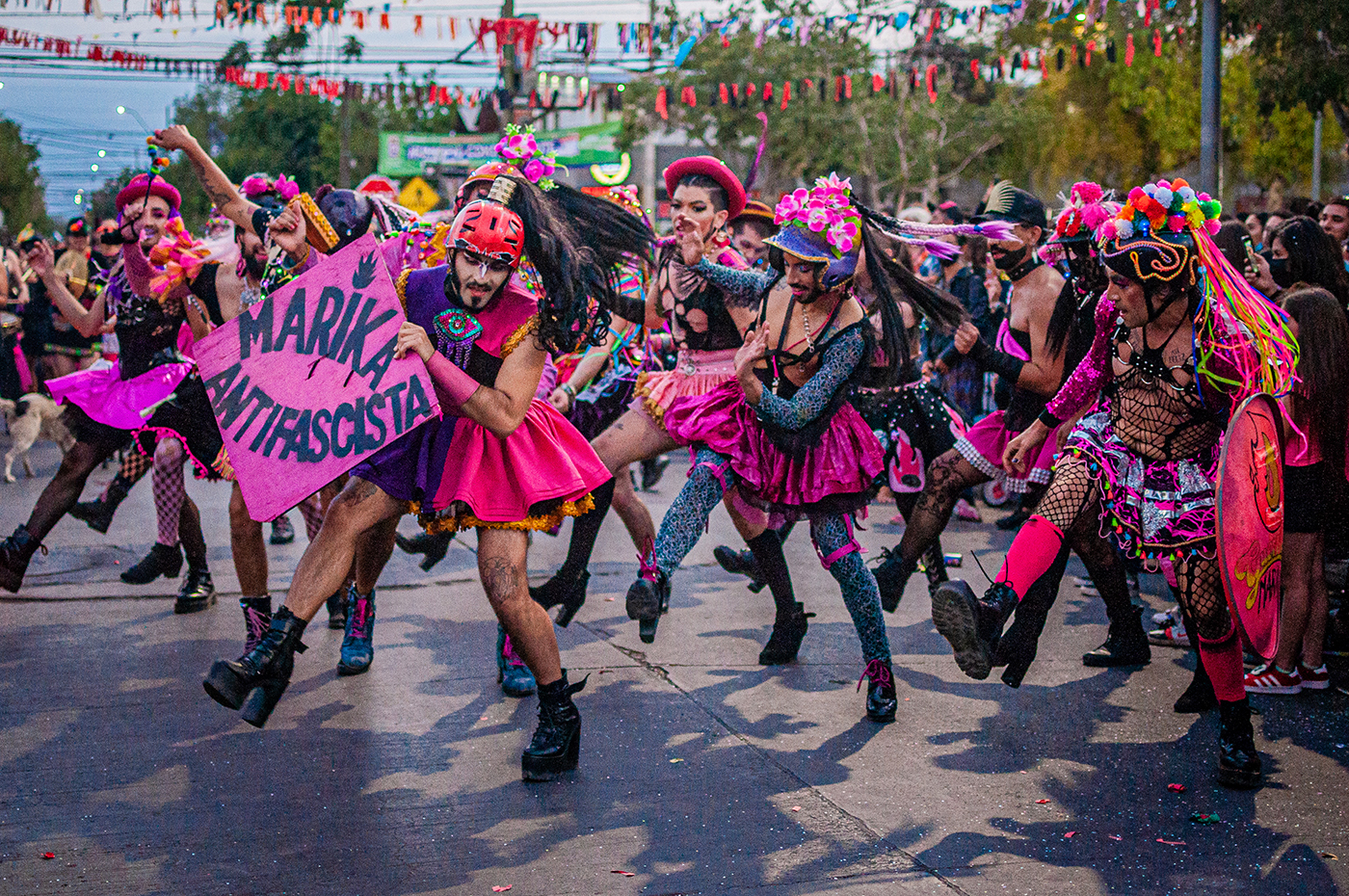 Ein Farbfoto zeigt eine tanzende Gruppe in schwarzen und pinkfarbenenen Netzkostümen mit kurzen Satinröcken und stilisierten Kopfbedeckungen auf einer festlich geschmückten Straße. Sie tragen ein Schild mit der Aufschrift 