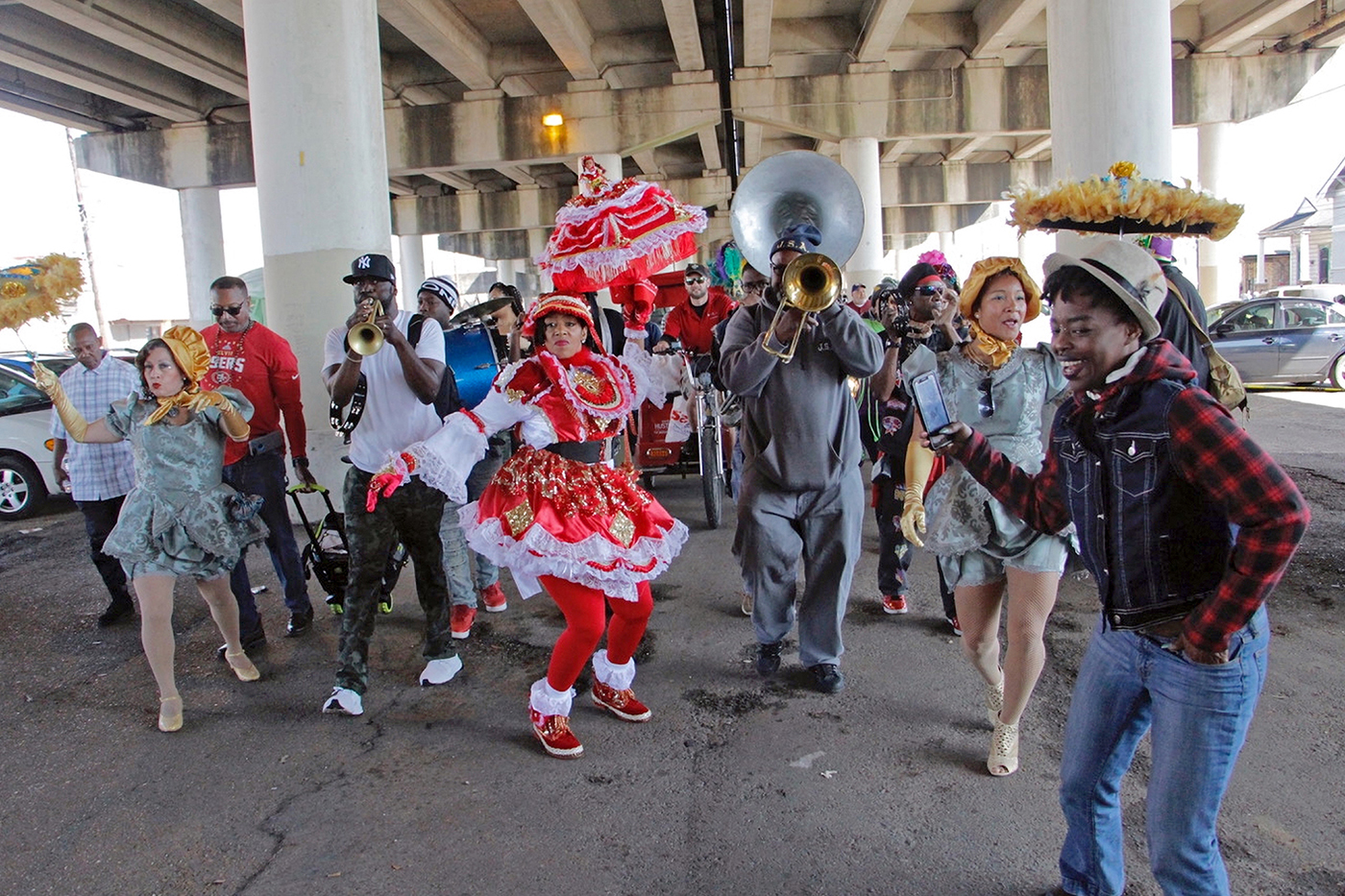 Ein Farbfoto zeigt eine tanzende, bunt kostümierte Gruppe in kurzen Rüschenkleidern und mit entsprechenden Kopfbedeckungen, die von einer Blaskapelle begleitet wird, unter einer Autobahnüberführung.
