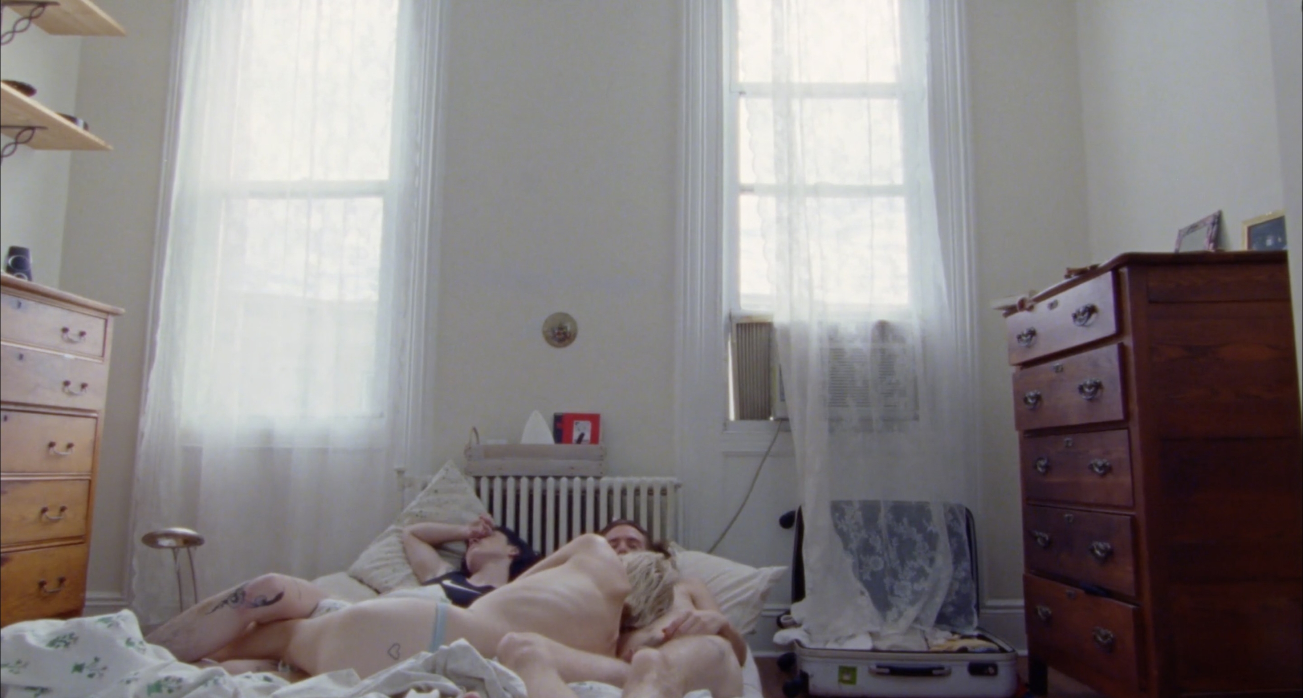 An image of three people in underwear, sleeping entwined on a mattress in the center of a small, sparsely furnished room. Soft daylight fills the space, filtered through nearly transparent lace curtains that are drawn closed.