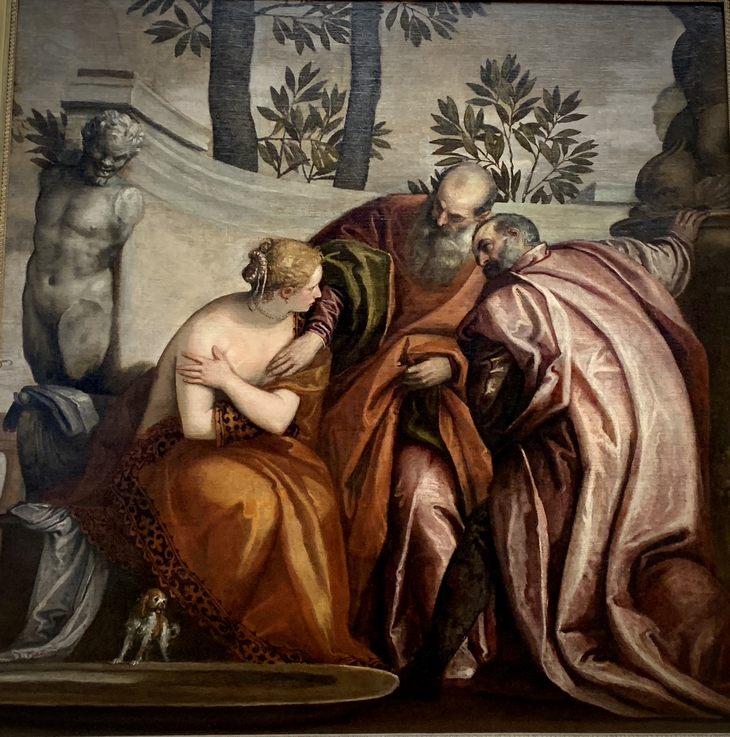 A Renaissance painting showing a partially draped woman seated beside two elderly men in rich robes, who lean toward her in an intense exchange. Behind them stands a classical statue and trees. The scene represents the biblical story of “Susanna and the Elders”, rendered with dramatic lighting and expressive gestures.