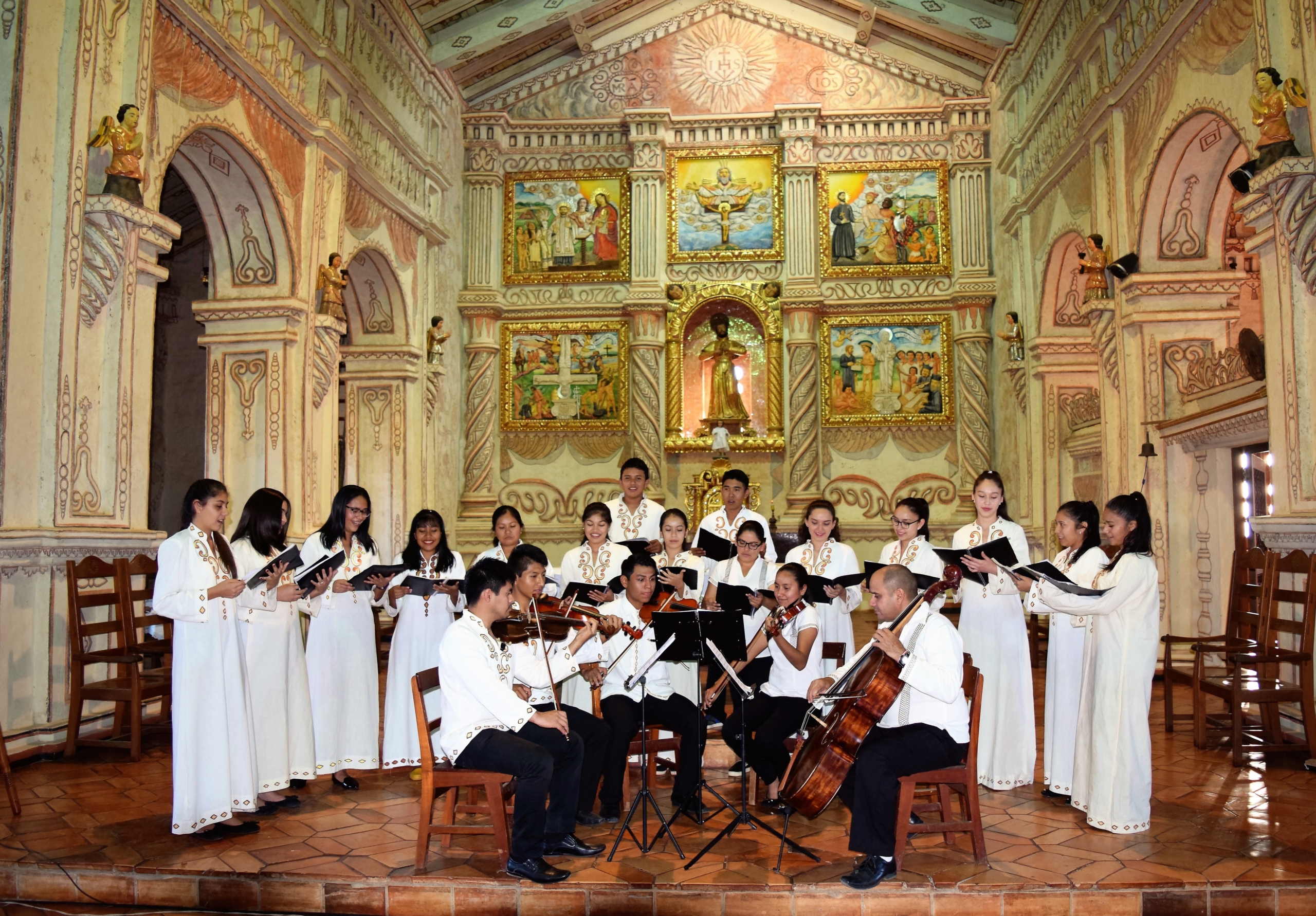 A photograph of people playing instruments and singing inside a highly ornate building. The interior features painted stucco walls with decorative arches and religious murals arranged behind an altar. In the center, there are five people seated playing string instruments (violin, viola and cello), dressed in white shirts and black pants. Behind them there is a group of 15 people singing, dressed in long white robes, all holding sheet music.