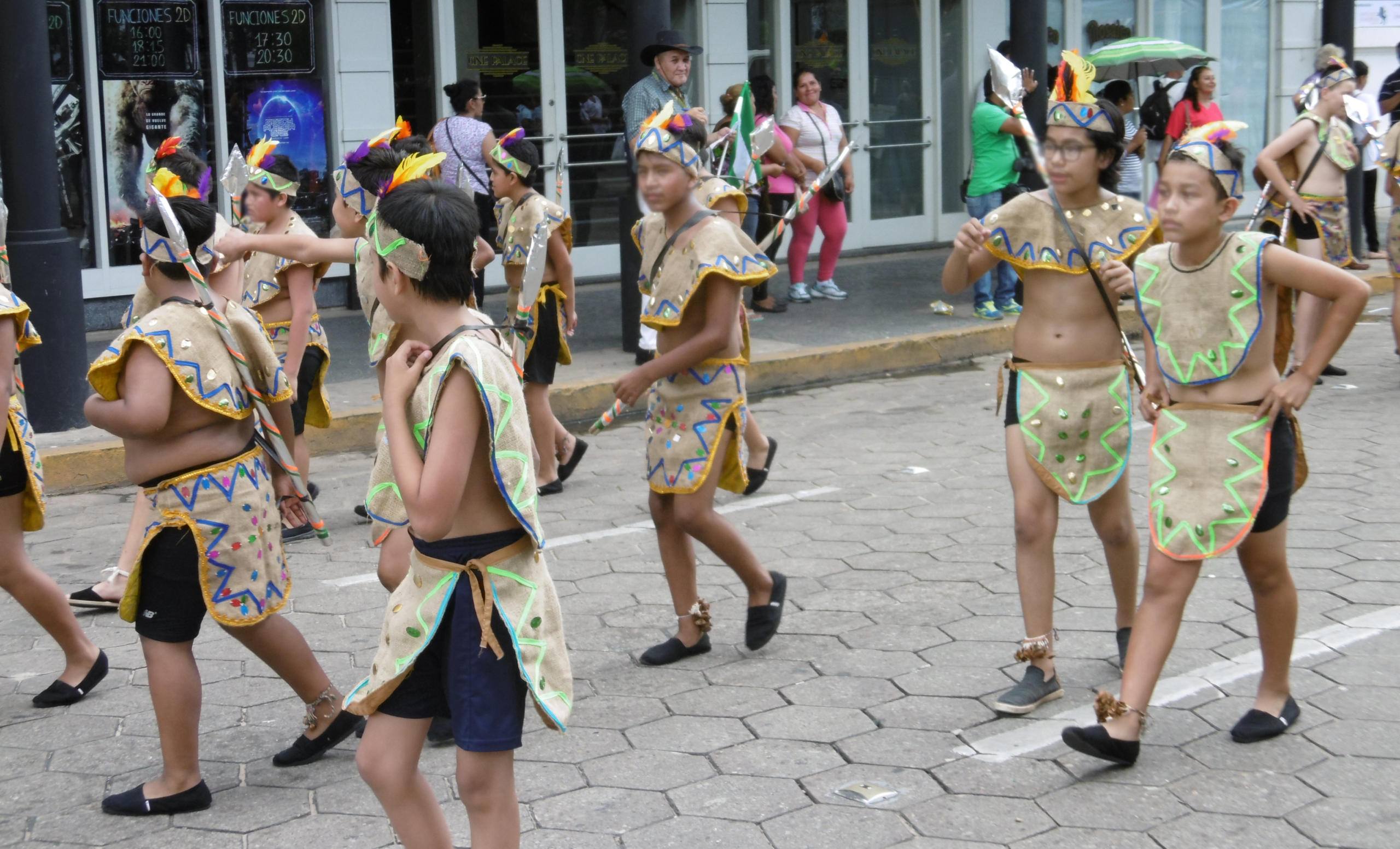A color photograph showing a group of children and young teenagers participating in a street parade or cultural performance. They are all wearing similar light-brown, simple costumes designed to resemble indigenous attire, consisting of a short tunic or cape over the shoulders and a loincloth or short skirt with geometric designs stitched in bright colors. They also wear headbands with feathers. The children are walking and dancing on a paved sidewalk/street. In the background, modern commercial buildings and adult onlookers can be seen, placing the event in a contemporary urban setting, likely in Latin America.