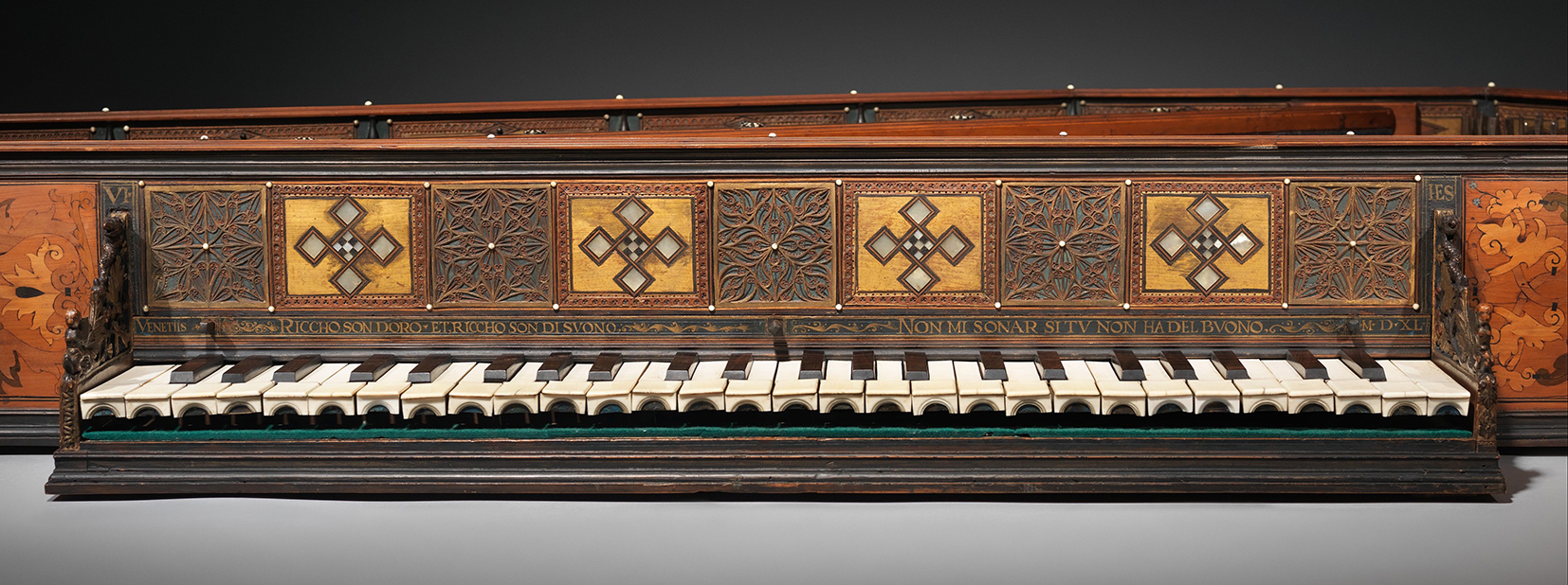 Close-up of an ornate virginal featuring intricately carved wooden panels and decorative inlays. The front panel displays alternating geometric and floral motifs with gold and dark wood accents, accompanied by an inscribed Italian phrase across the front. The white ivory keys and dark wooden sharps are framed by detailed craftsmanship, with floral marquetry on the instrument’s sides. The design exemplifies Venetian artistry and luxury from the 16th century.
