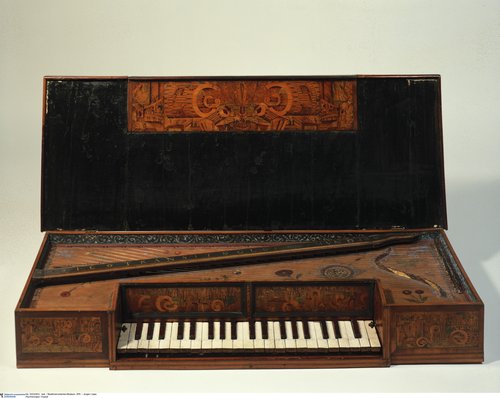 A virginal with its lid open, showcasing an elegant wooden case decorated with intricate marquetry and carved floral patterns. The keyboard features white and dark keys set into a rectangular frame, while the soundboard displays delicate painted and inlaid details, including ornamental rosettes. The instrument’s lid interior bears decorative wood inlay in symmetrical designs, highlighting early Baroque craftsmanship.