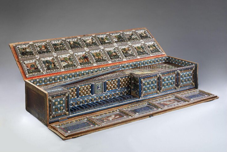 Exquisitely decorated virginal with its lid open, revealing elaborate Renaissance-era craftsmanship. The instrument’s interior is richly adorned with colorful geometric patterns, gilded accents, and miniature painted panels depicting architectural scenes and figures. The keyboard and surrounding frame feature intricate blue, gold, and white inlay designs, emphasizing luxury and artistic detail. The open lid displays a series of framed paintings bordered by ornate motifs, showcasing both musical and visual artistry.
