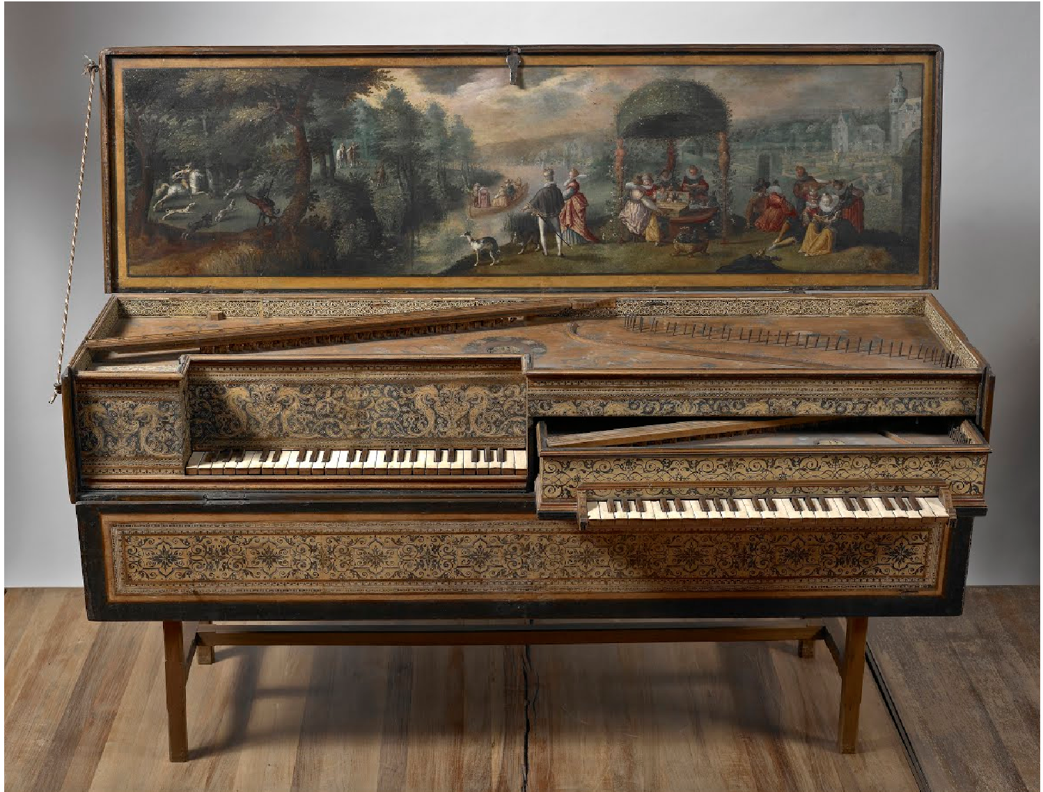 A double-manual virginal with elaborately decorated casing and an open lid featuring a pastoral landscape painting. The artwork inside the lid depicts elegantly dressed figures in a garden setting, engaged in music and conversation beneath a domed arbor, with a distant cityscape and lush trees in the background. The harpsichord’s exterior showcases intricate floral and scrollwork patterns in gold and black, emphasizing fine craftsmanship and Baroque elegance. Two tiers of ivory and ebony keys extend from the richly ornamented body, resting on wooden legs