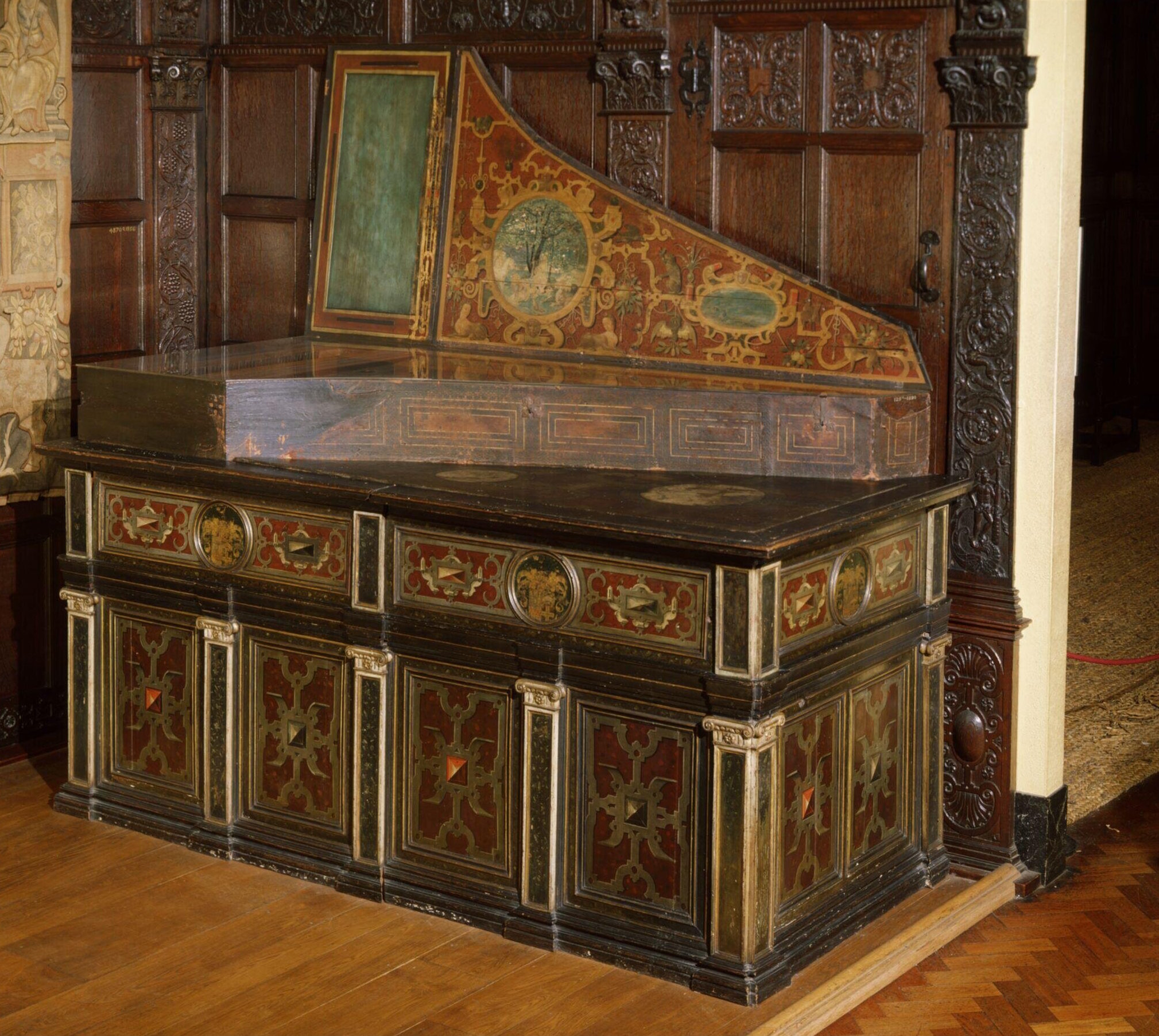 Claviorgan integrated into an ornate wooden cabinet, displayed in an elaborately paneled interior. The instrument features richly decorated panels in red, green, and gold with geometric and heraldic motifs framed by gilded accents. The open lid reveals painted scenes with intricate scrollwork and a central medallion depicting a pastoral landscape. The instrument’s base includes detailed carved columns and inlaid designs, blending fine furniture craftsmanship with musical artistry typical of 16th–17th century design.