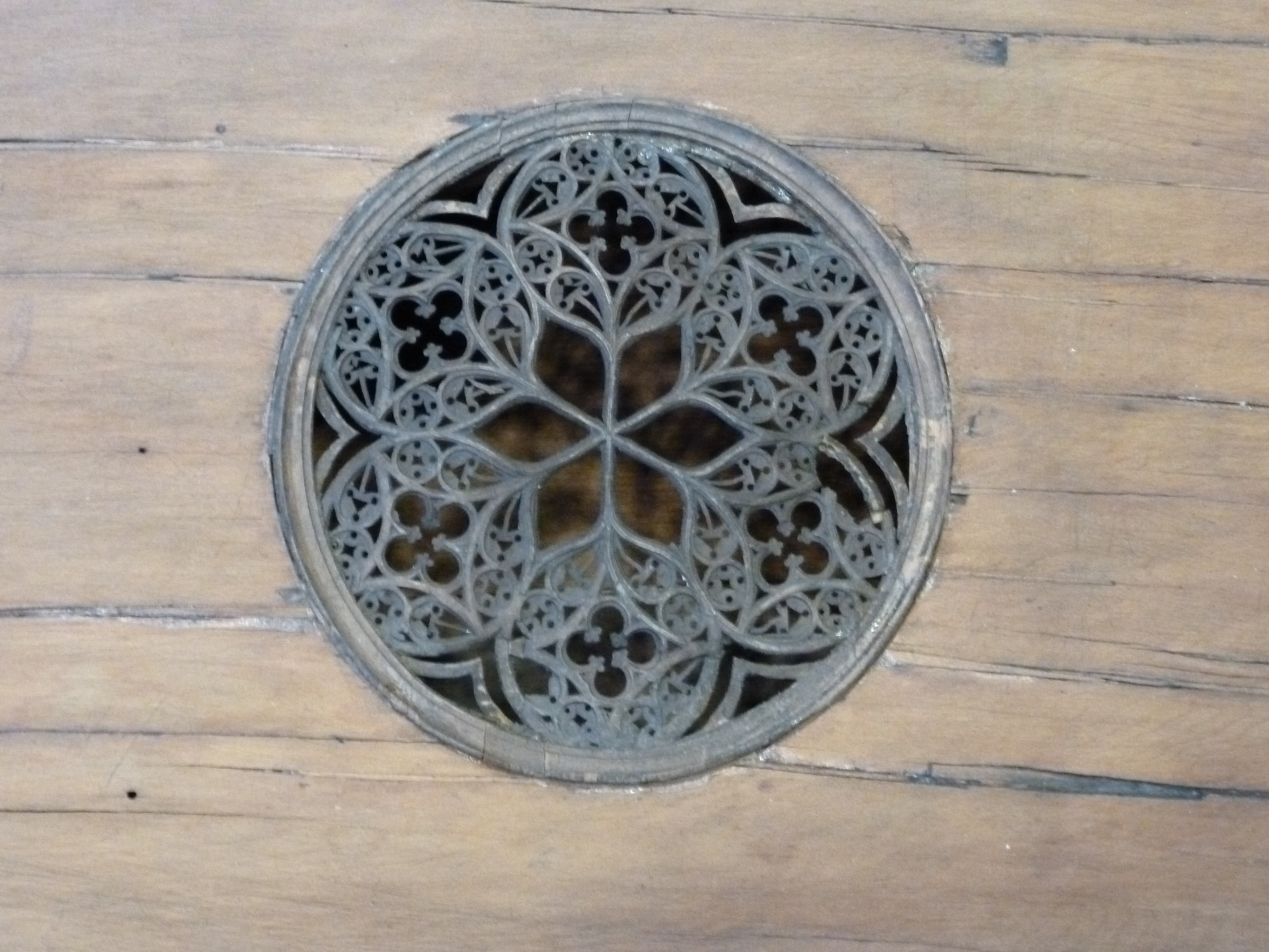 Close-up of an ornate circular sound hole rosette carved into the wooden soundboard of an antique harpsichord. The intricate Gothic-style design features interlacing floral and geometric patterns forming a symmetrical star or flower motif. The finely detailed latticework contrasts with the smooth, aged wood surrounding it, highlighting expert craftsmanship and historical artistry.