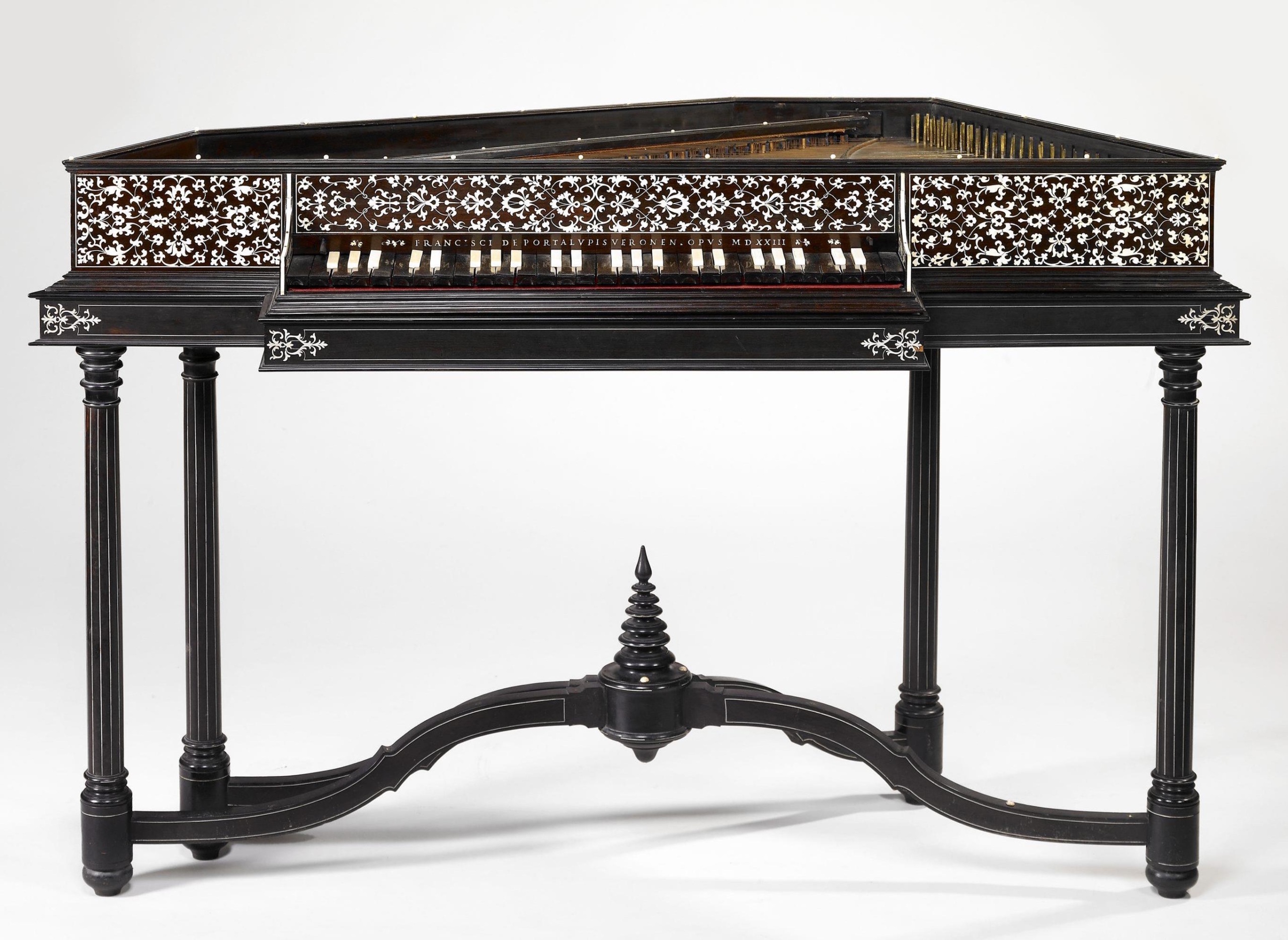Antique Italian harpsichord with intricate floral ivory inlay and dark wood finish, standing on four fluted legs connected by a carved stretcher. The body features ornate white vine patterns along the sides and front, with a keyboard of alternating white and dark keys. The front panel bears an inscription reading “Francisci de Portalupis Veronensis Opus MDXXIII,” indicating it was crafted in 1523. This Renaissance-era musical instrument exemplifies fine craftsmanship and decorative artistry.