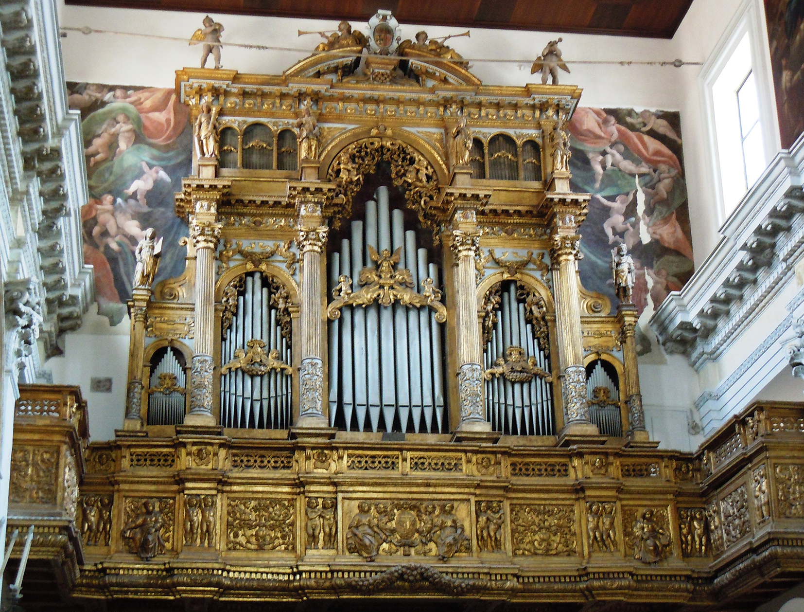 The grand organ in the Church of Monteoliveto, featuring an opulent gilded wooden facade richly carved with Renaissance motifs. The organ case is divided into three main sections with ornate columns, statues, and intricate reliefs, framing the silver organ pipes. Behind it, a vivid fresco decorates the upper wall, enhancing the architectural splendour of the church interior.