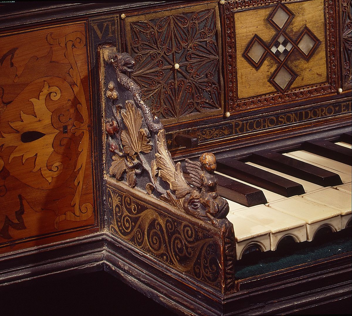 Close-up of an ornate antique virginal, featuring richly carved and gilded wood decorations. The image shows detailed floral and scrollwork carvings, a small sculpted figure, and inlaid geometric patterns. The keys are made of ivory and dark wood, and part of an inscription in gold lettering is visible above them.