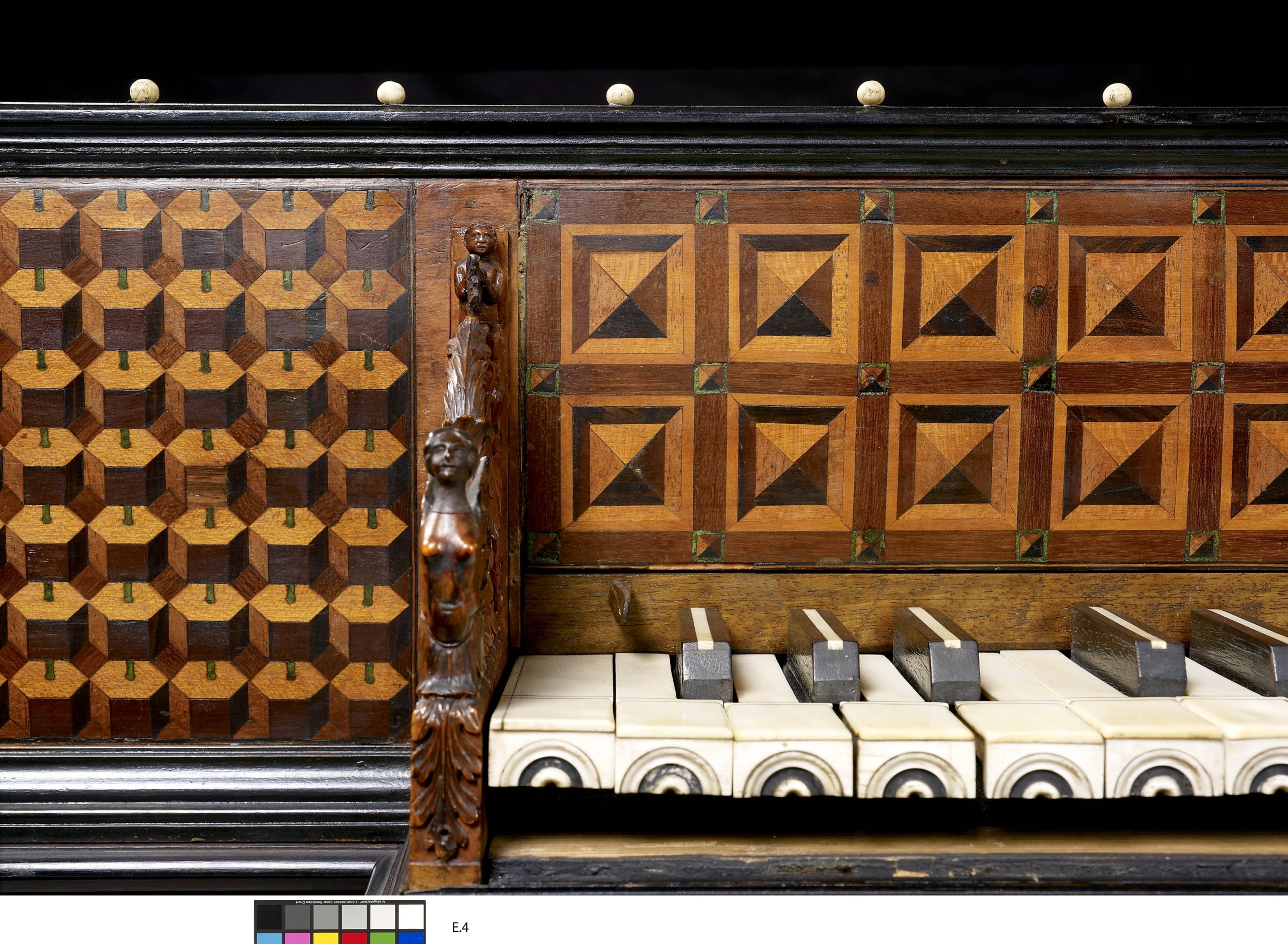 Close-up of an ornate antique virginal, featuring intricate wood inlay patterns and detailed carvings. The left panel displays a geometric cube motif, while the right panel shows diamond-shaped square designs. Between them is a carved wooden figure with a human bust. The ivory and black keys are visible at the bottom, and a black frame with decorative knobs runs along the top edge.