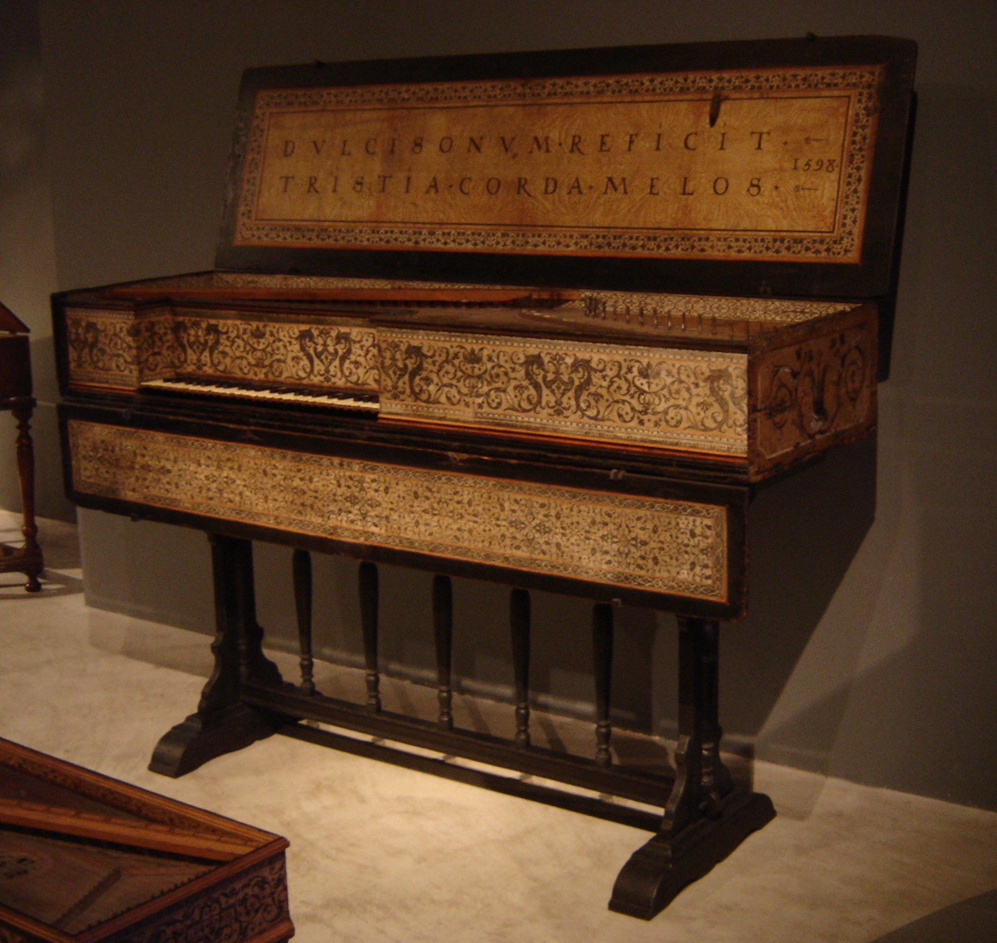 A richly decorated antique virginal in a museum setting. The instrument features intricate floral and geometric patterns in black and gold across its body and lid. The lid bears a Latin inscription reading “DVLCISONVM REFICIT TRISTIA CORDA MELOS” with the date 1598. The harpsichord stands on a dark wooden base with turned supports, set against a neutral wall and floor.