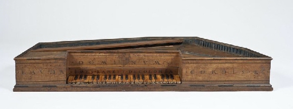 Antique polygonal virginal, made of varnished wood with a single manual keyboard featuring alternating light and dark keys. The rectangular and slightly trapezoidal case has Latin inscriptions engraved on the front panel. It misses a lid and thus reveals the inner strings. The design is simple yet elegant, showcasing Renaissance craftsmanship typical of 16th- to 17th-century instruments