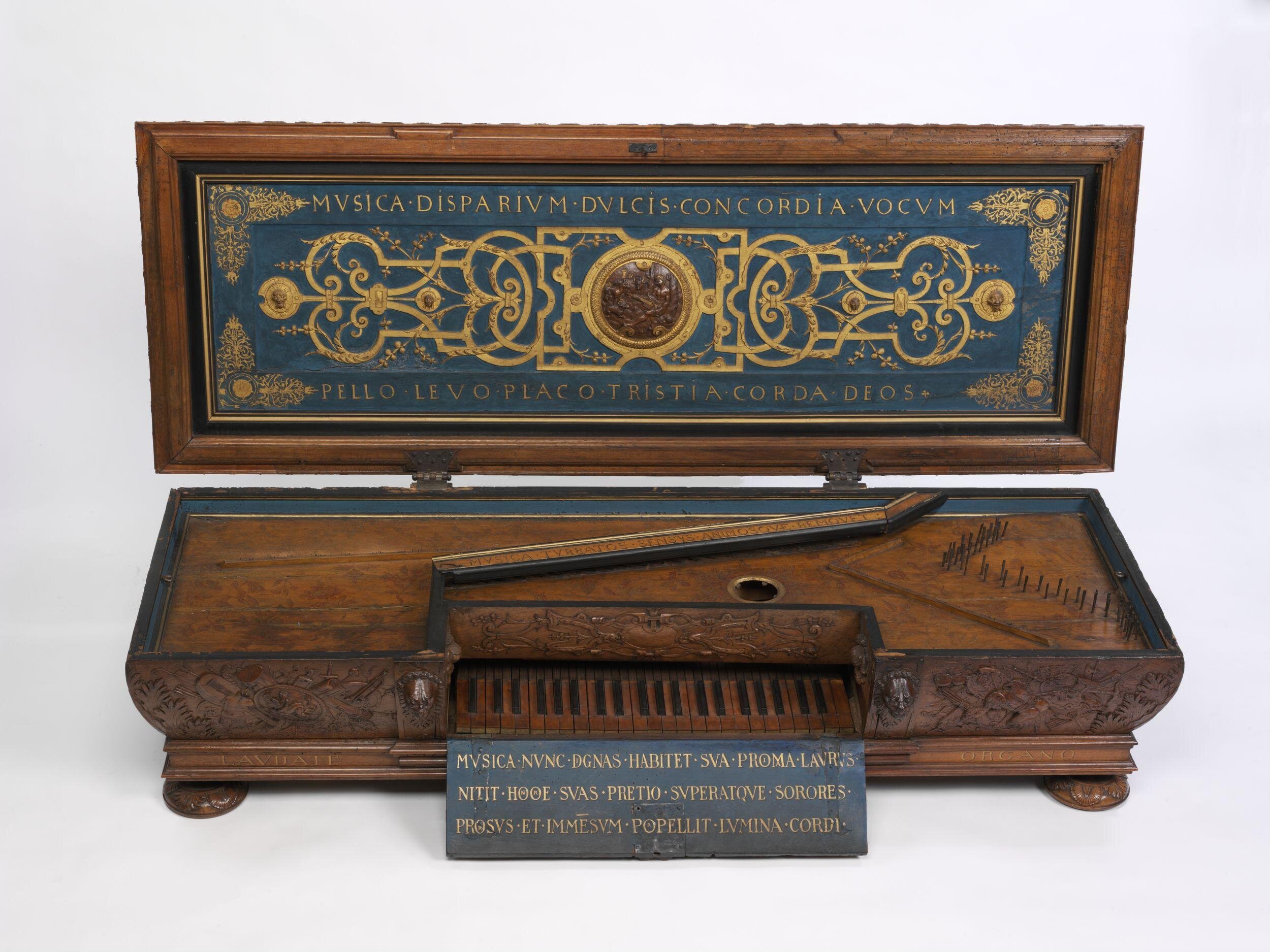 Renaissance-era virginal crafted from carved wood with elaborate decorative details and Latin inscriptions. The instrument features a rectangular case with a single manual keyboard of alternating light and dark keys, surrounded by ornate carvings of foliage and mythological motifs. The open lid reveals an intricate blue and gold painted interior with symmetrical scrollwork, gilded ornamentation, and the Latin phrase “MVSICA DISPARIVM DVLCIS CONCORDIA VOCVM.”