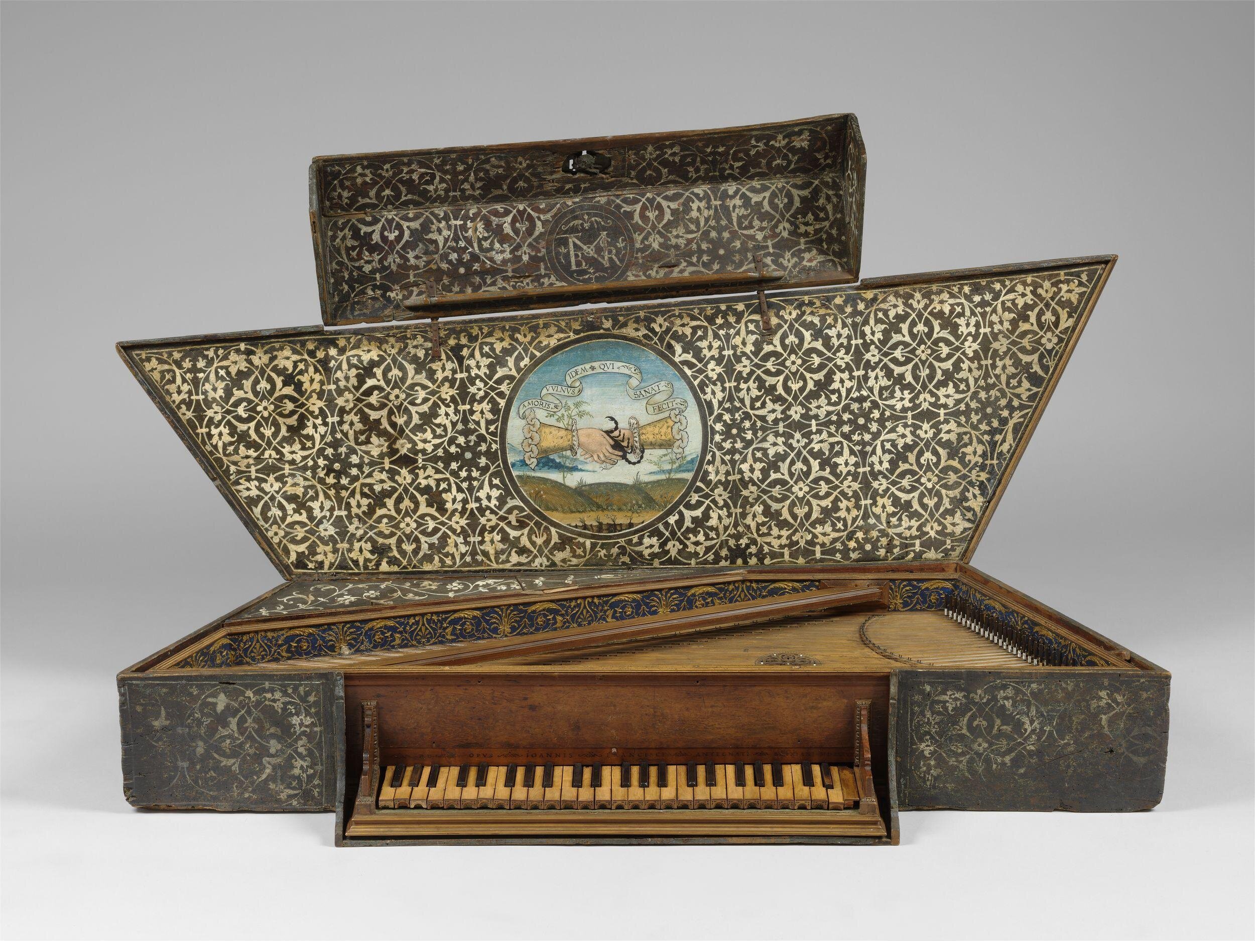 An ornate early virginal, with its lid open to reveal an intricate interior. The lid is decorated with a detailed floral pattern and a central painted medallion showing two hands clasping over a landscape, with Latin text on a scroll. The wooden keys and strings of the instrument are visible, surrounded by decorative motifs in gold and blue.