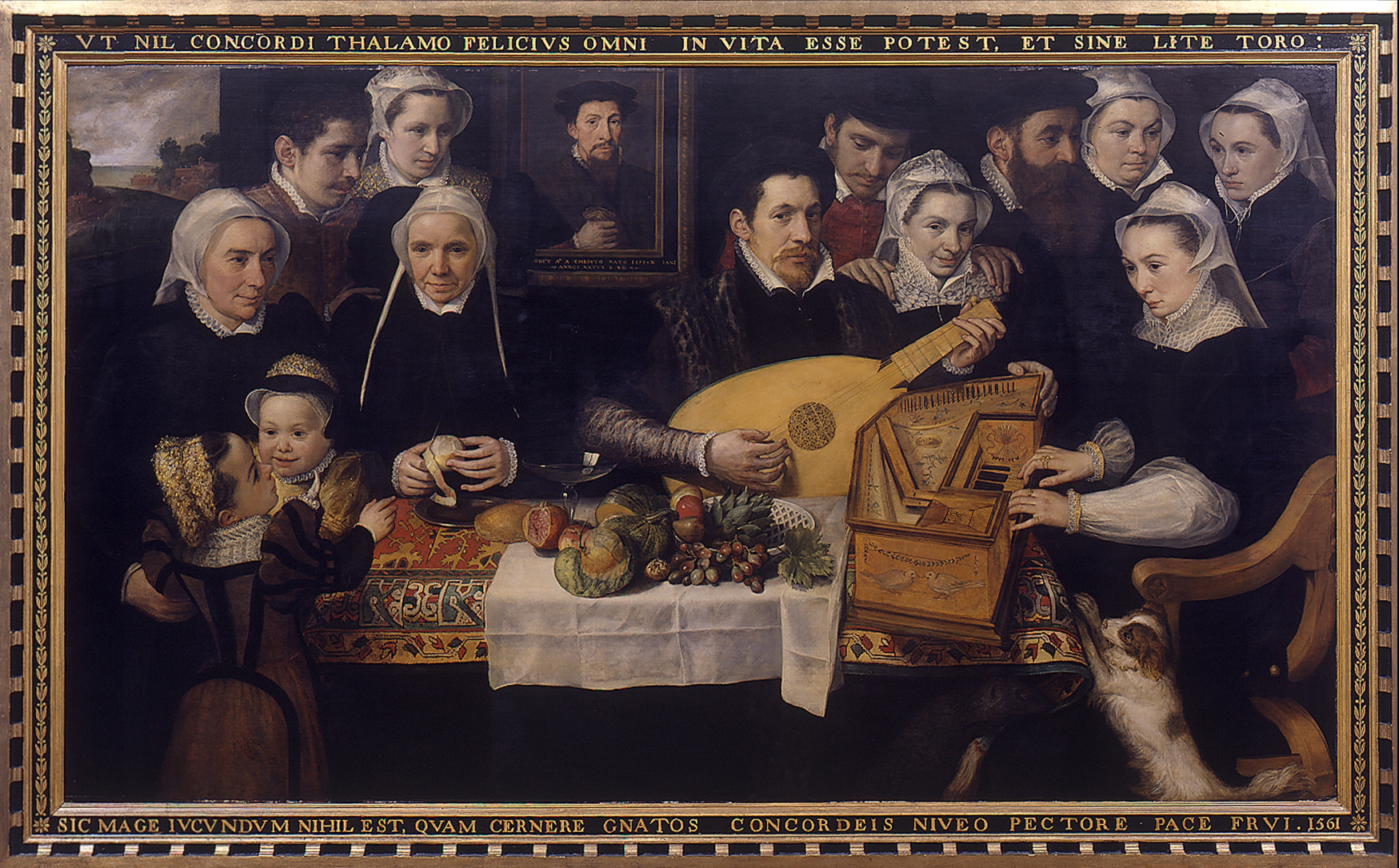 Renaissance family portrait showing a large group gathered around a table covered with fruit and bread. The central male figure plays a lute while a woman beside him performs on a virginal. The family members, dressed in rich 16th-century attire with lace collars and dark garments, include men, women, and children, all wearing white coifs or bonnets. A small dog stands at the right, gazing upward. The Latin inscription above and below the painting praises marital and familial concord.