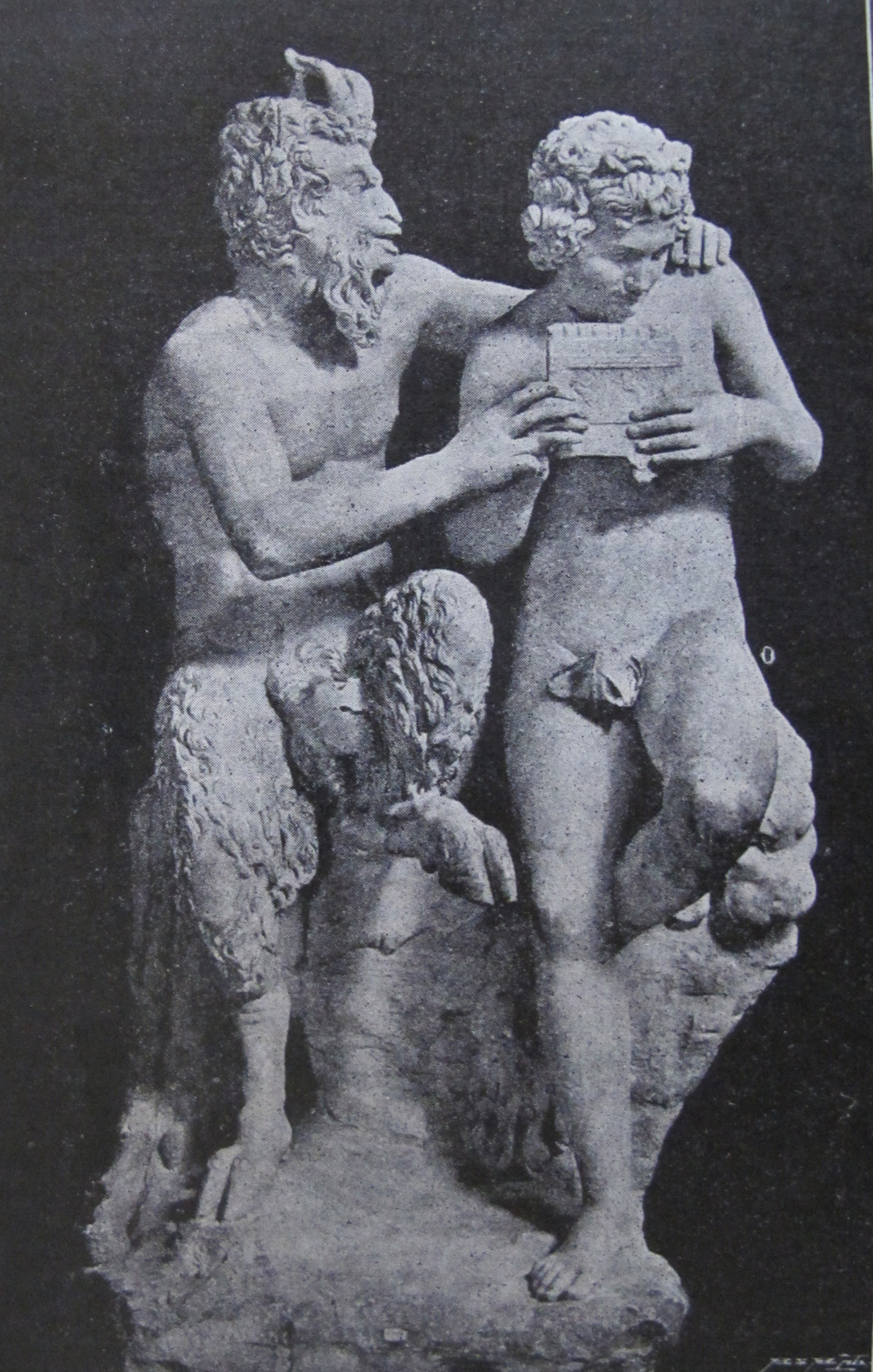 A classical marble sculpture depicting the god Pan teaching the young shepherd Daphnis to play the panpipes. Pan, identifiable by his goat legs, horns, and beard, sits on a rock with one arm resting affectionately on Daphnis’s shoulder, guiding his hands on the instrument. Daphnis stands beside him, gazing intently at the pipes. The figures are rendered with detailed anatomical precision and naturalistic expression.