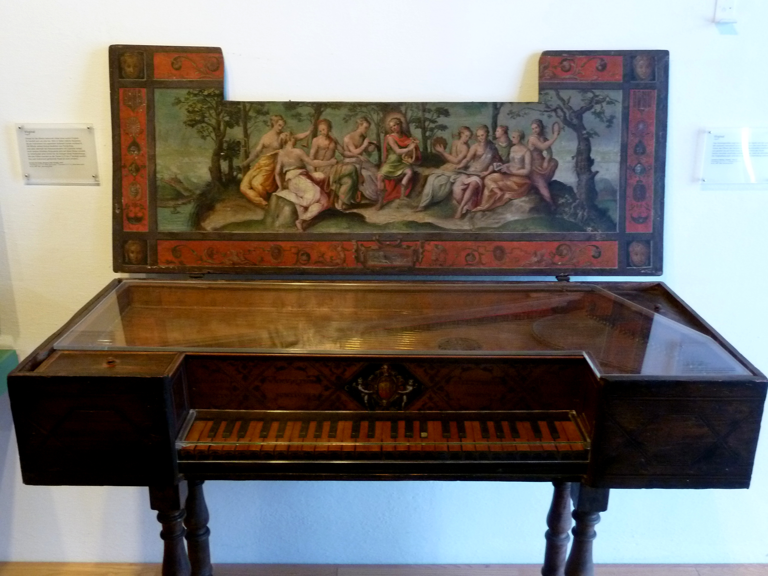 A virginal, displayed in a museum setting. The open lid reveals a painted panel depicting a mythological or allegorical scene of nine muses gathered outdoors, each holding musical instruments or scrolls. The frame of the lid is richly decorated with red and black ornamental motifs. The keyboard below has wooden keys, and the instrument’s body features intricate marquetry and craftsmanship.