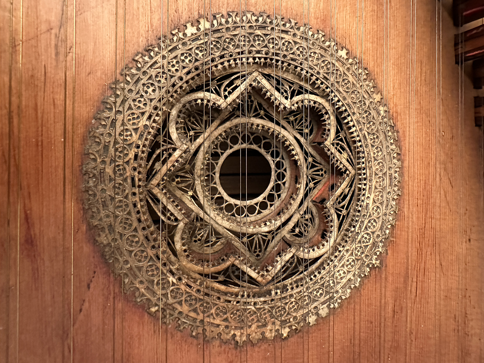Close-up of an ornate circular sound hole rosette on a virginal. The intricate design features interlaced geometric and floral patterns carved in delicate detail, below metal strings running vertically across the image.