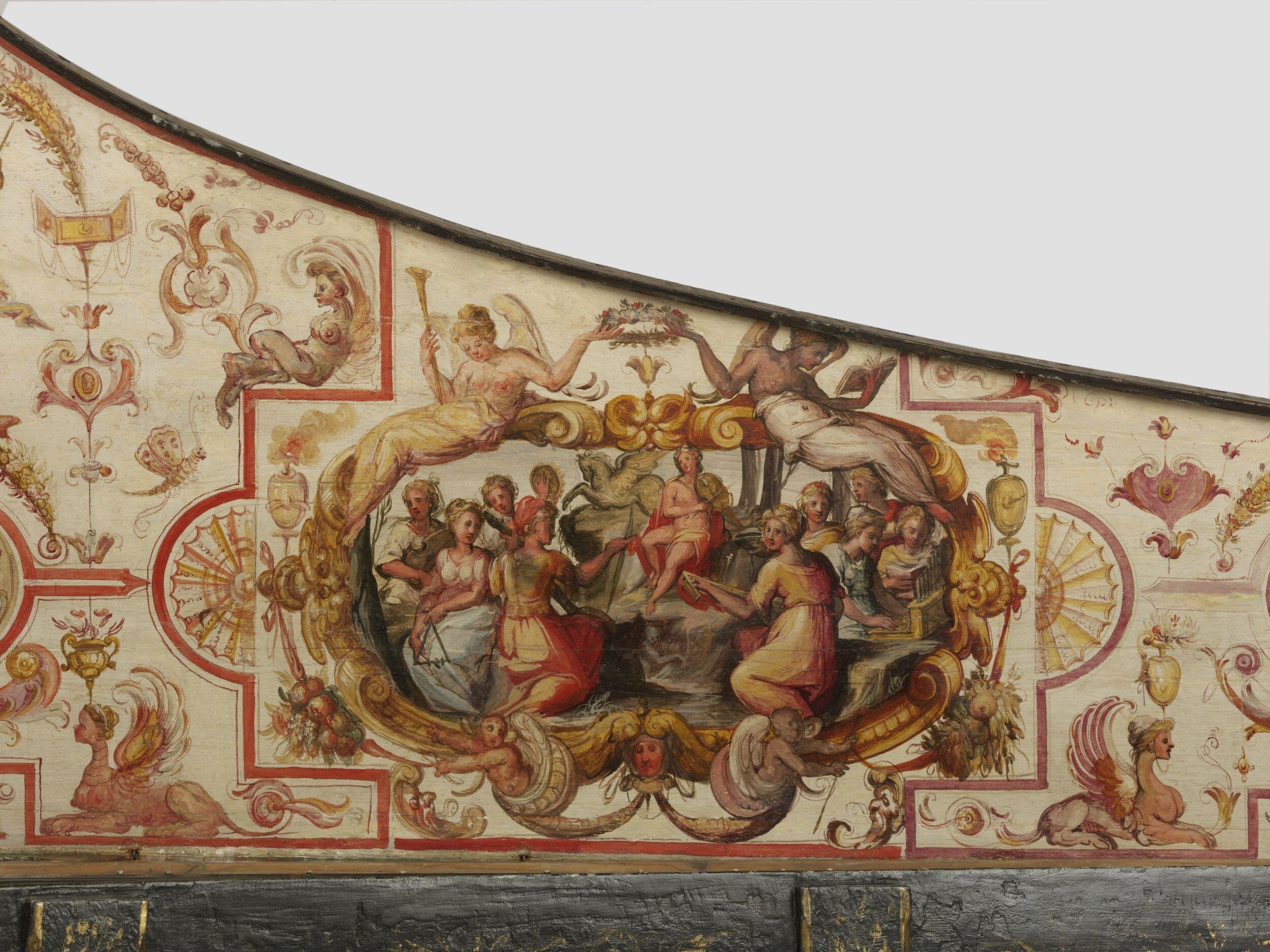 A richly decorated panel from a harpsichord, featuring an elaborate painted scene surrounded by ornate designs. The central image depicts a group of classical figures, representing the Muses and Apollo, engaged in music and conversation, with instruments such as a lyre and an organ. Two winged allegorical figures recline above, holding a laurel wreath and a book. The surrounding decoration includes intricate scrollwork, masks, sphinxes, and floral motifs rendered in red, gold, and cream tones.