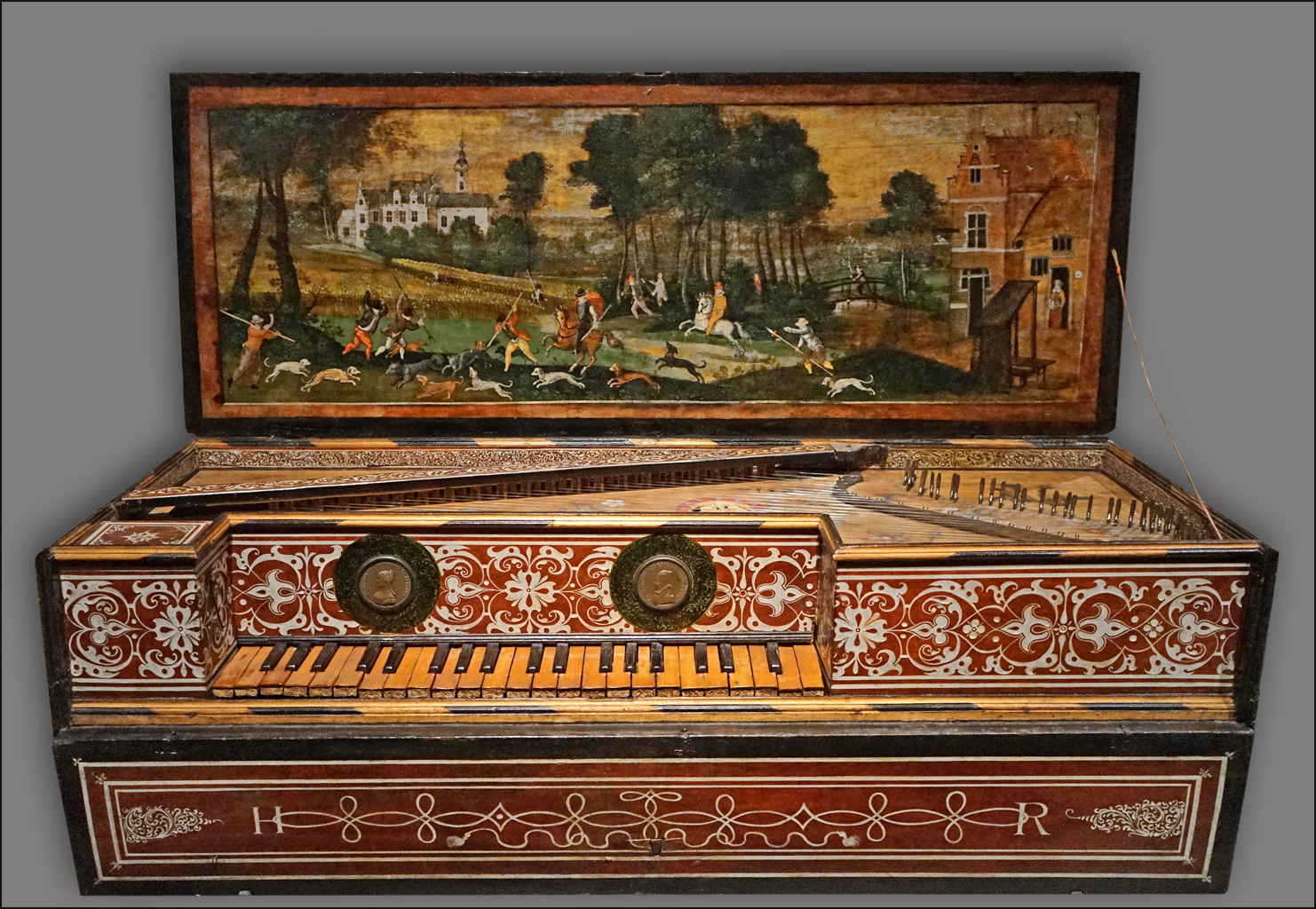 A richly decorated virginal with intricate red, white, and gold floral designs on its case and keyboard area. The open lid features a painted pastoral scene depicting a lively hunting party with riders on horseback, dogs chasing game, and a distant manor house surrounded by trees. The instrument’s craftsmanship and detailed artwork exemplify the opulence and artistic refinement of early modern European musical instruments, blending visual art with musical function.