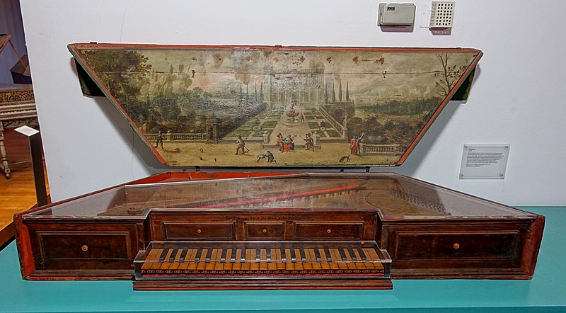 A historical virginal displayed in a museum, featuring a richly painted interior lid. The artwork depicts a formal garden scene with fountains, pathways, and elegantly dressed figures strolling or conversing among manicured trees and architectural structures. The wooden instrument has a warm brown finish with a simple, elegant keyboard and decorative panels.