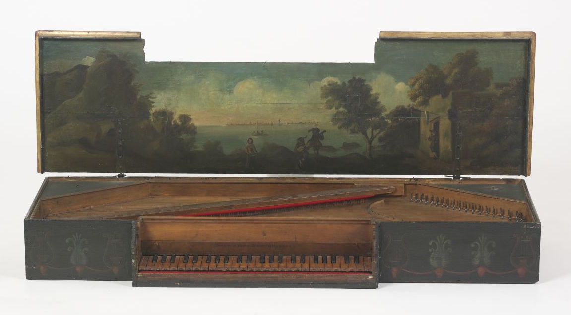 An antique virginal with its lid open, revealing a painted interior scene of a serene landscape. The artwork depicts figures in the foreground beside trees, with a distant view of a calm body of water and a city skyline under a softly clouded sky. The harpsichord’s wooden case is dark with simple floral motifs, and its natural wood keyboard and red felt lining add warmth to the design.