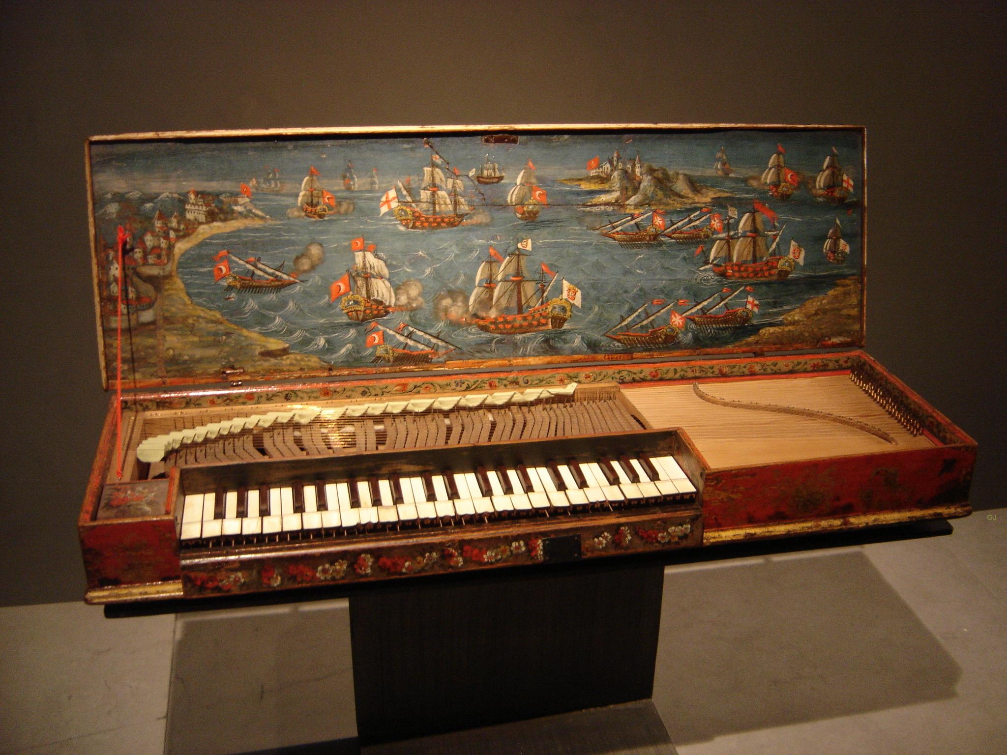 A historical clavichord with its lid open, revealing a vibrant painted scene of a naval battle. The artwork depicts numerous sailing ships engaged in combat, with billowing sails, cannon fire, and flags flying over turbulent blue waters near a rocky coastline. The instrument’s exterior is decorated with floral motifs in red and gold, while the keyboard and inner mechanics are exposed, showcasing its intricate craftsmanship.