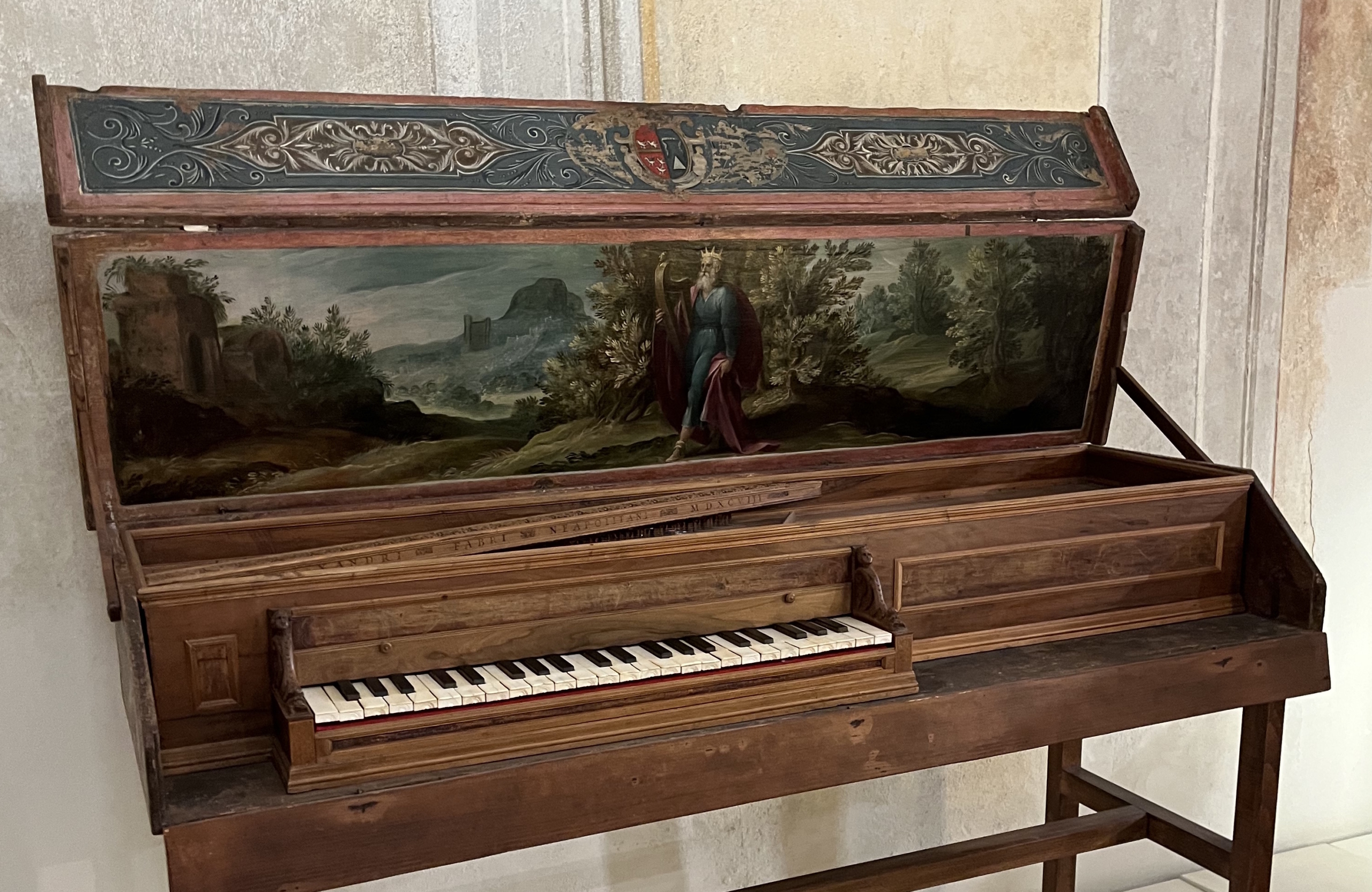 A wooden keyboard instrument with its lid open, revealing a painted interior panel. The artwork inside depicts a regal, crowned figure (King David) in a flowing robe holding a harp, standing in a lush landscape with trees, rolling hills, and distant ruins. The instrument’s exterior features ornate carved and painted decoration, including a heraldic crest on the upper lid.