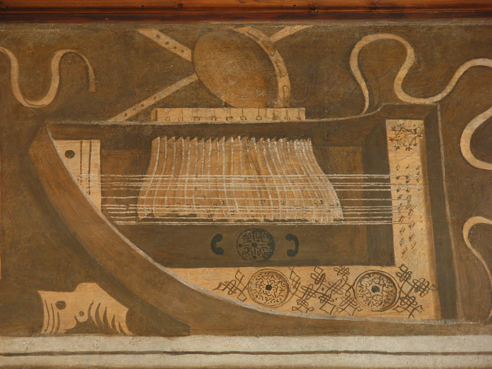 A wall painting depicting a stringed keyboard instrument, shown in stylized geometric and earthy tones of brown and beige. The instrument has multiple strings stretched across a rectangular wooden frame, with decorative patterns, circular motifs, and flowing ribbon-like shapes around it.