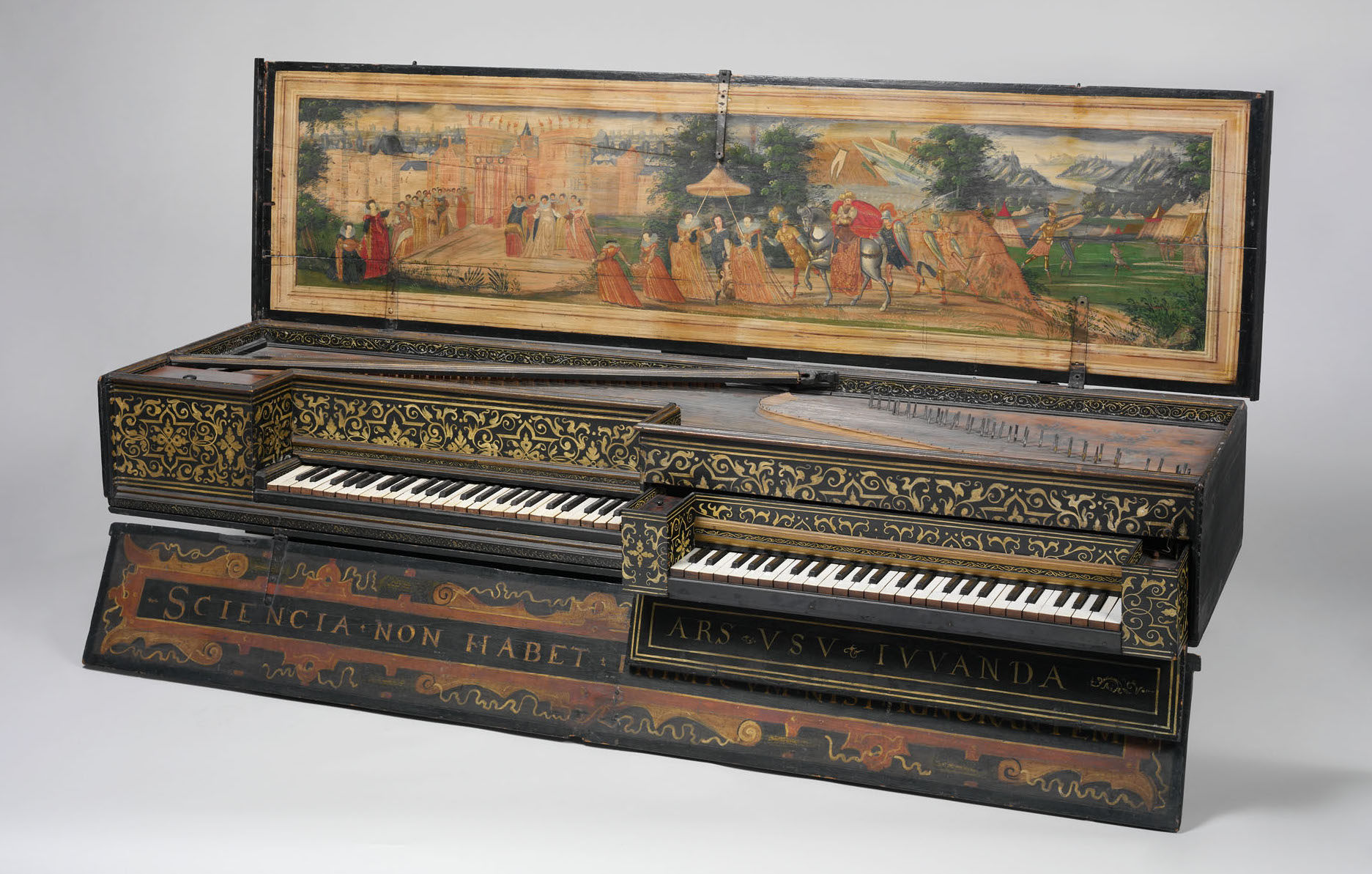 A richly decorated Renaissance harpsichord with intricate gold and black patterns on its case and two keyboards. The open lid reveals a painted scene depicting a procession: elegantly dressed figures, some on horseback and others on foot, approach a castle gate, while mountains and tents appear in the background. The Latin inscription “SCIENTIA NON HABET INIMICUM NISI IGNORANTEM” adorns the lower panel, meaning “Knowledge has no enemy but the ignorant.”