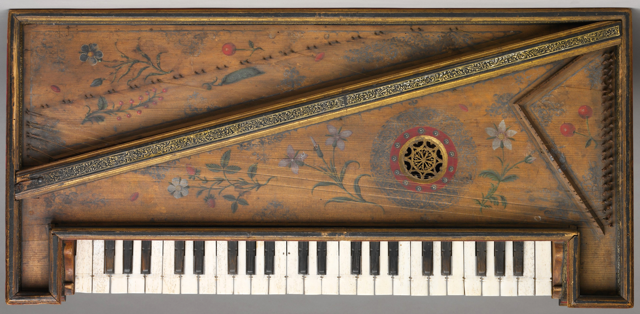 Overhead view of a Renaissance virginal, a rectangular keyboard instrument with a wooden case decorated with floral motifs. The soundboard features painted flowers and leaves in muted tones of red, green, and white, along with a circular, intricately carved soundhole rose accented by a red ring. The keyboard has alternating white and black keys, and the instrument’s structure includes ornate gold-trimmed borders.